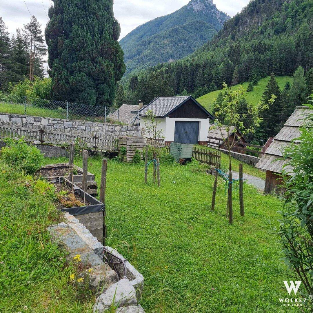 Weitläufiger Garten mit Rasenfläche und jungen Bäumen, im Hintergrund ein kleines Gebäude und Berge.