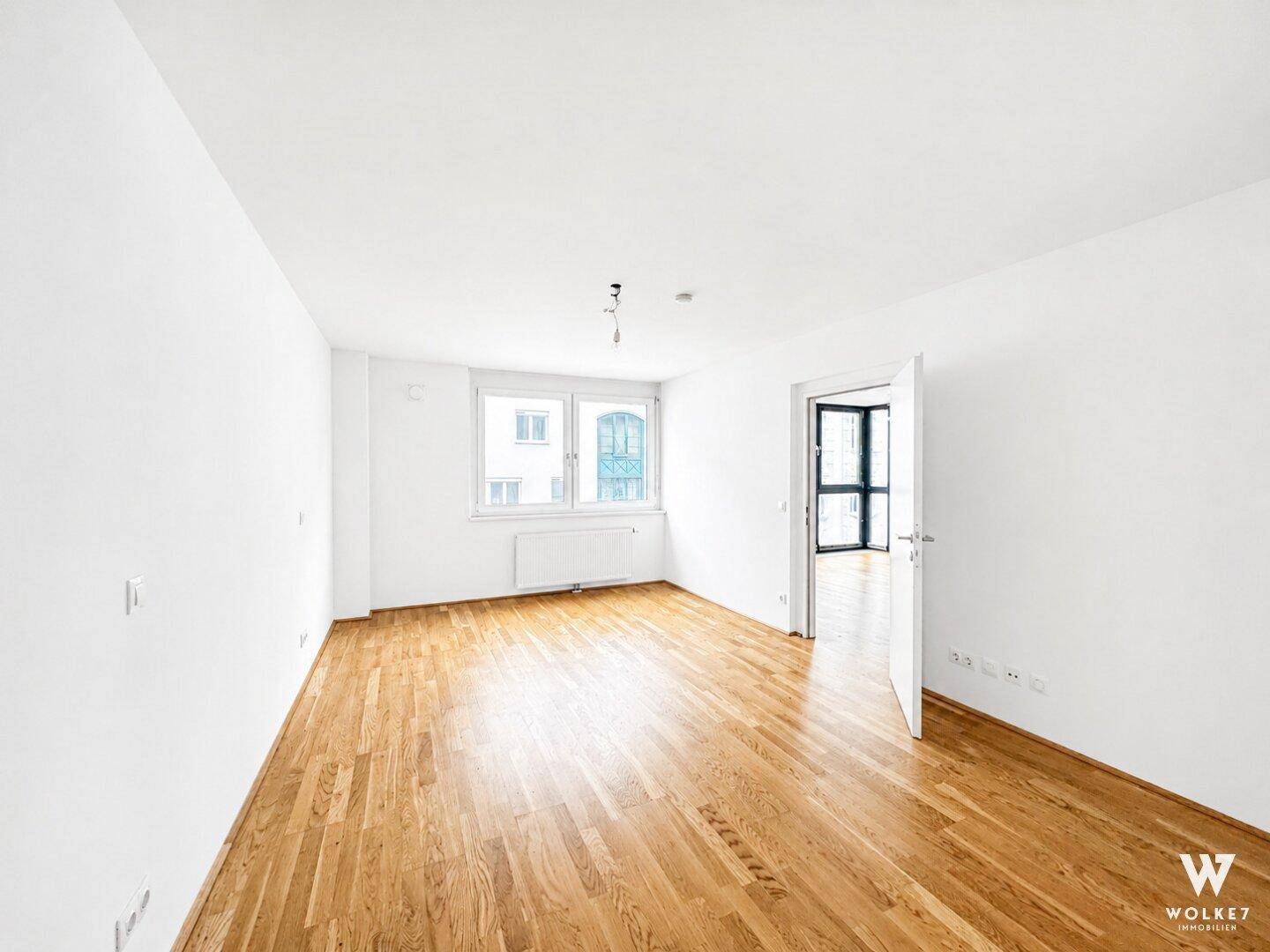 Leerer Raum mit hellem Parkettboden, weißen Wänden und einem Fenster.