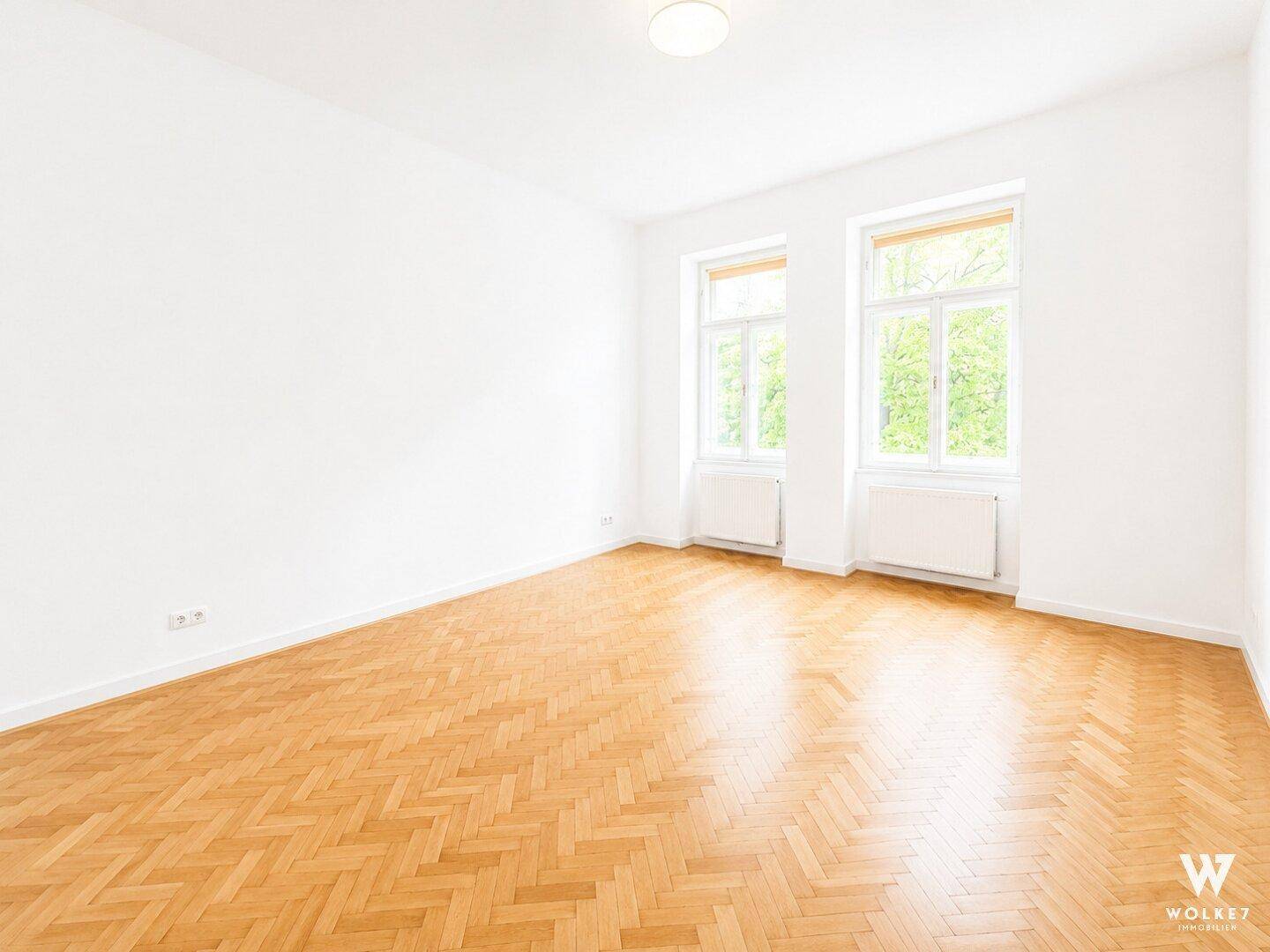 Heller, leerer Raum mit klassischem Parkettboden und zwei großen Fenstern.