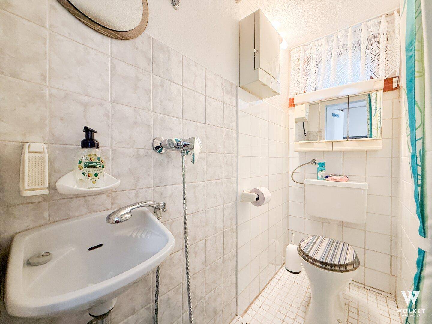 Kleines Badezimmer mit weißen Fliesen, Waschbecken, Toilette und einem Fenster.