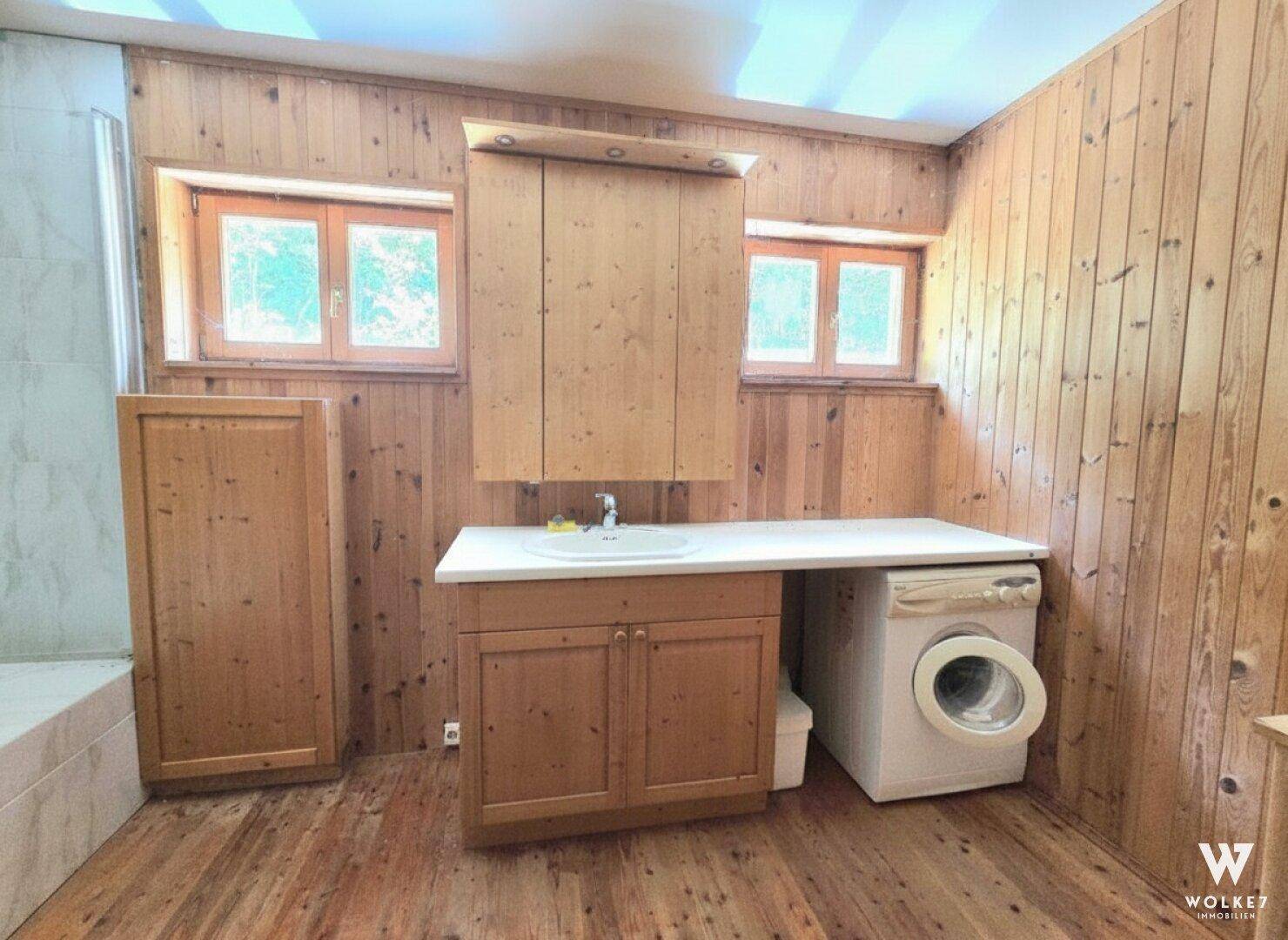 Badezimmer mit Holzwänden, Einzelwaschbecken, Waschmaschine und zwei Fenstern.