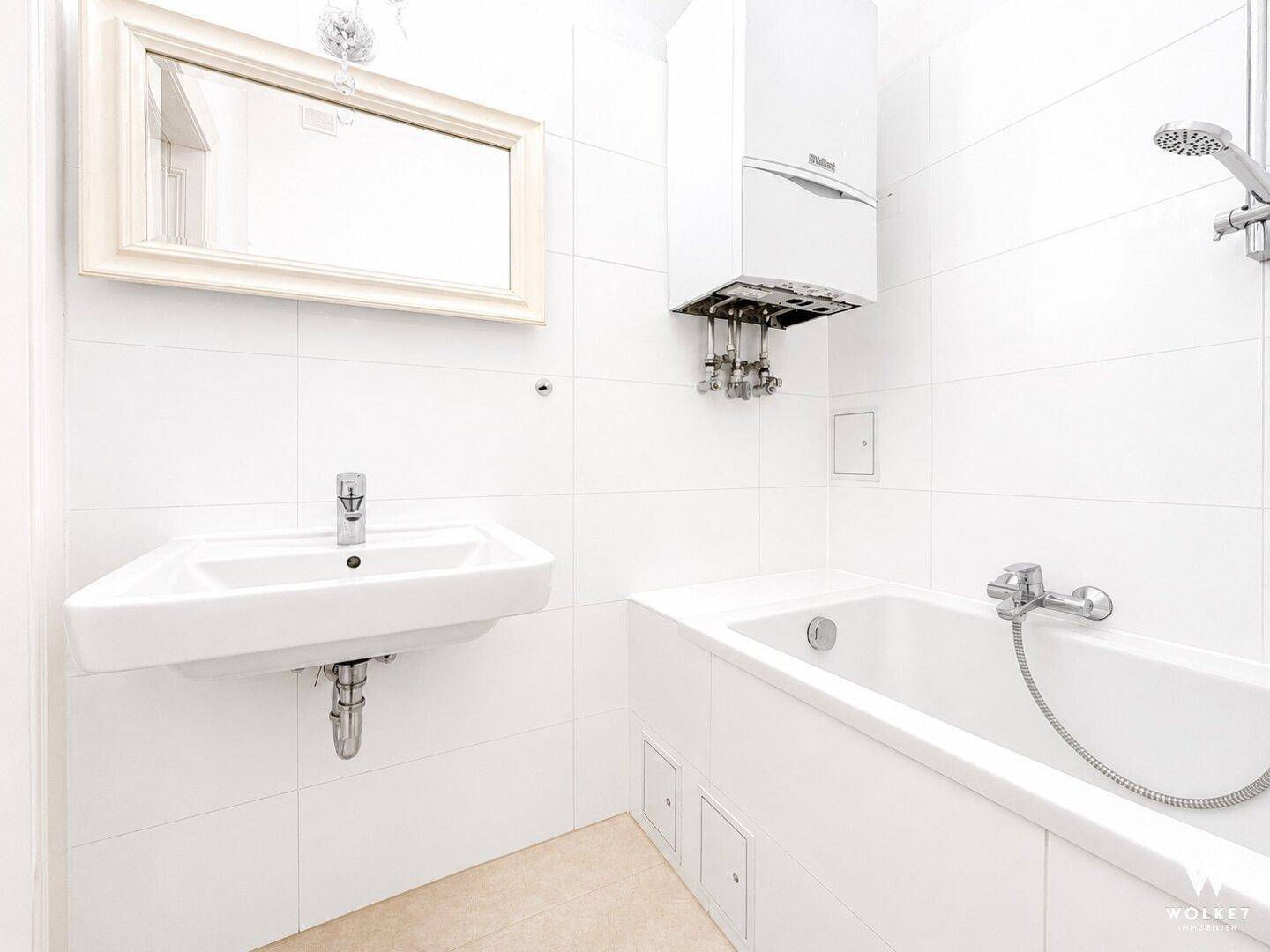 Helles Badezimmer mit weißen Fliesen, Badewanne, Waschbecken und Gastherme an der Wand.