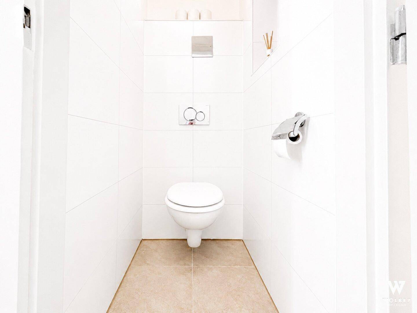 Separates WC mit wandhängender Toilette und weißen Fliesen an Wänden und Boden.