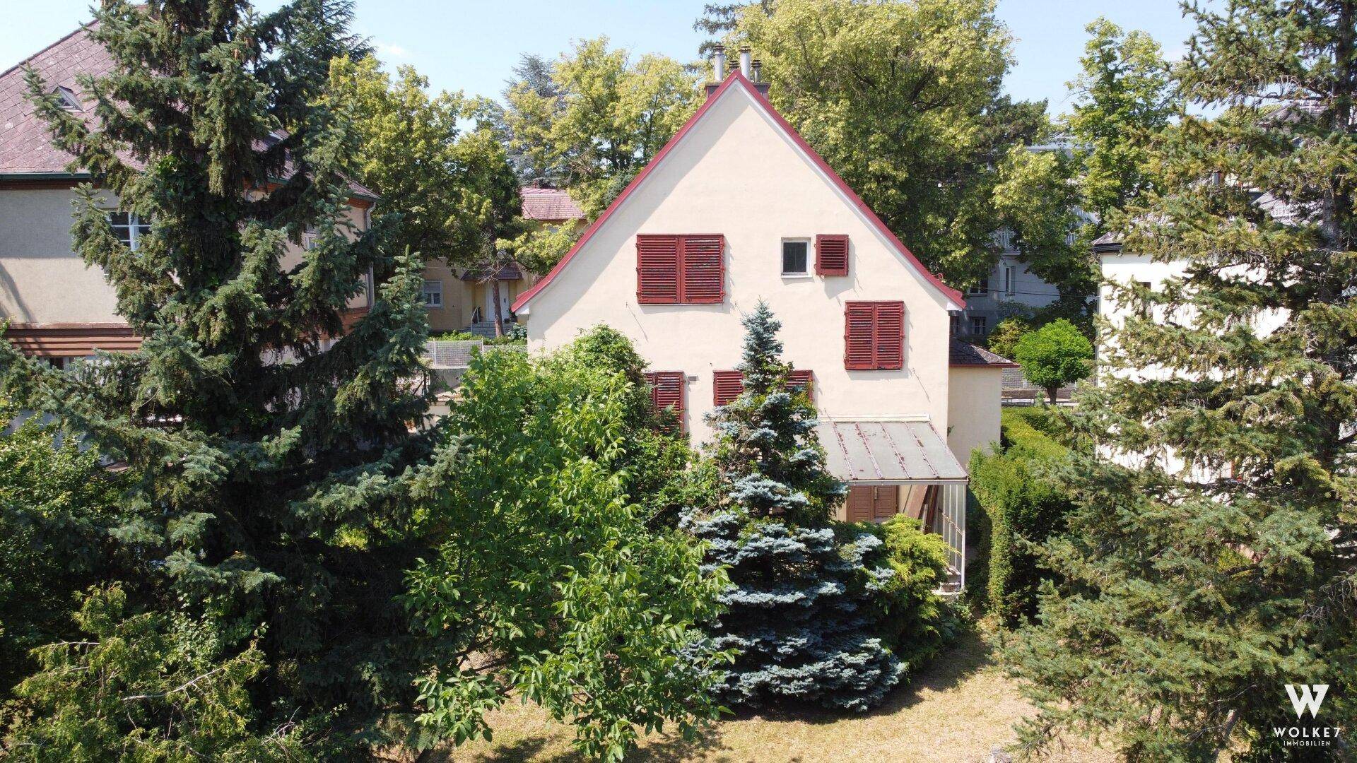 Vorderansicht des Hauses mit roten Fensterläden, umgeben von hohen Nadelbäumen und einer grünen Rasenfläche.