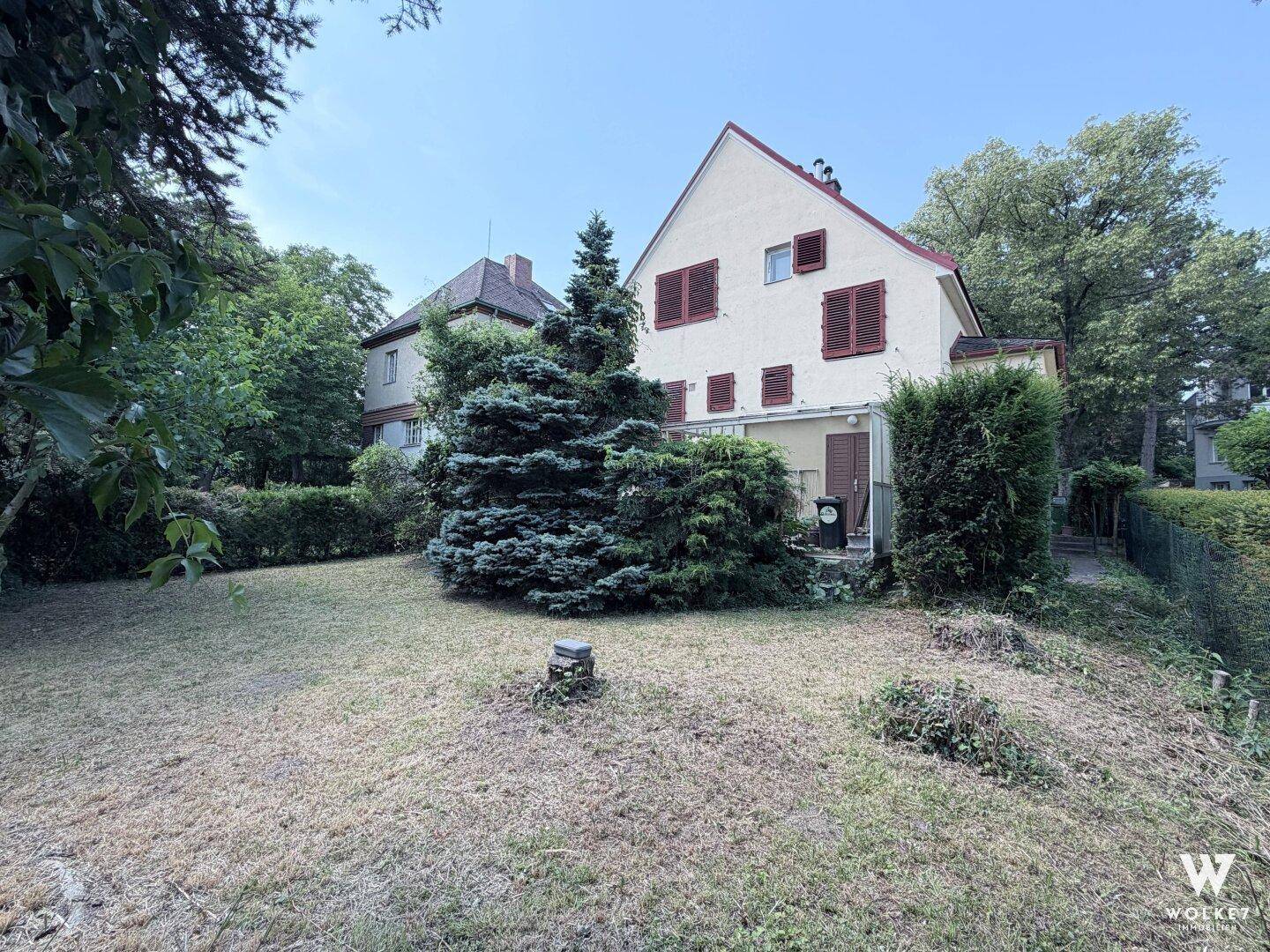 Blick vom Garten auf die Rückseite des Hauses mit einer kleinen überdachten Terrasse und viel Grünfläche.