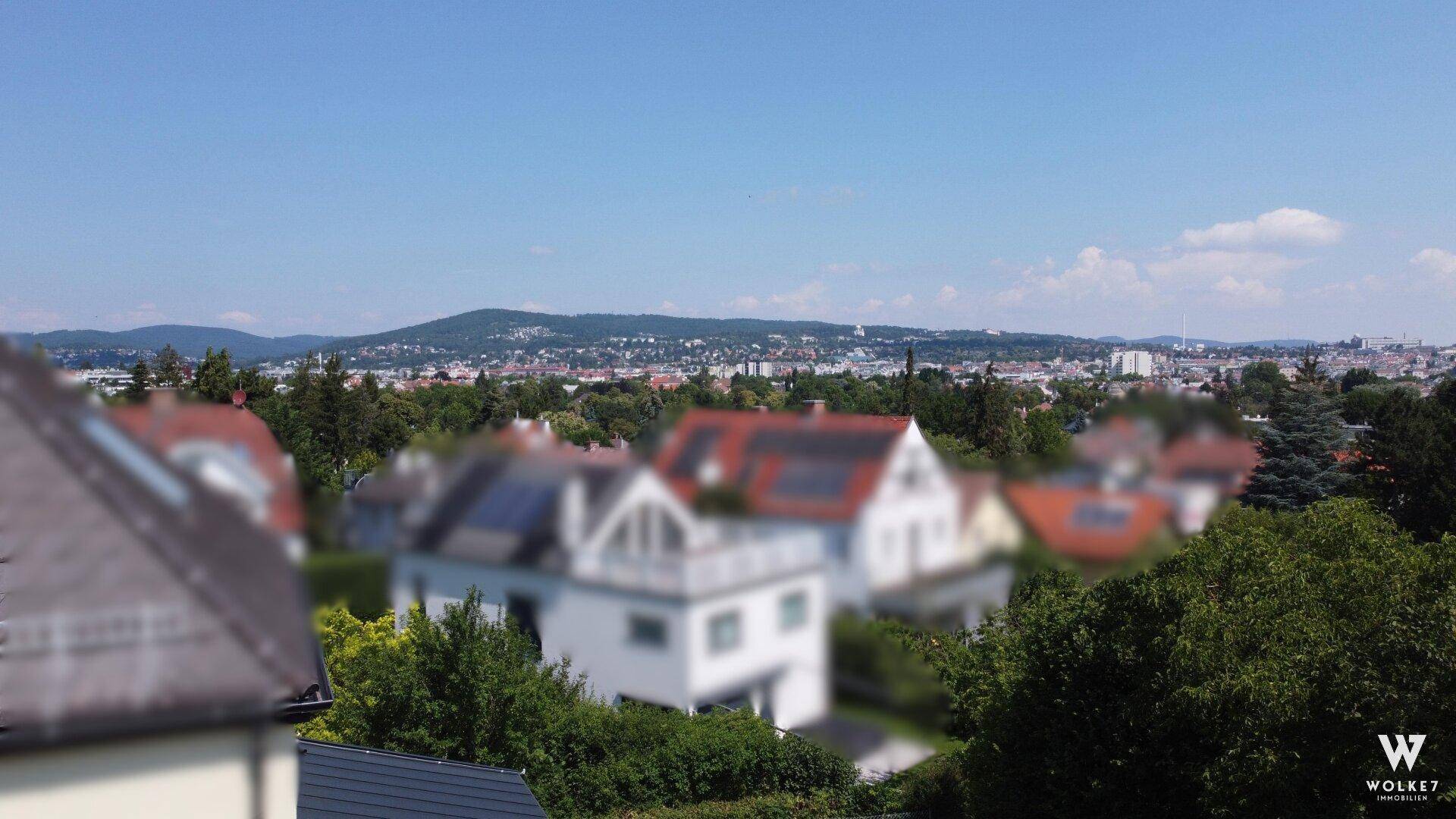 Weitblick über eine Stadtlandschaft mit vielen Dächern und bewaldeten Hügeln im Hintergrund unter blauem Himmel.