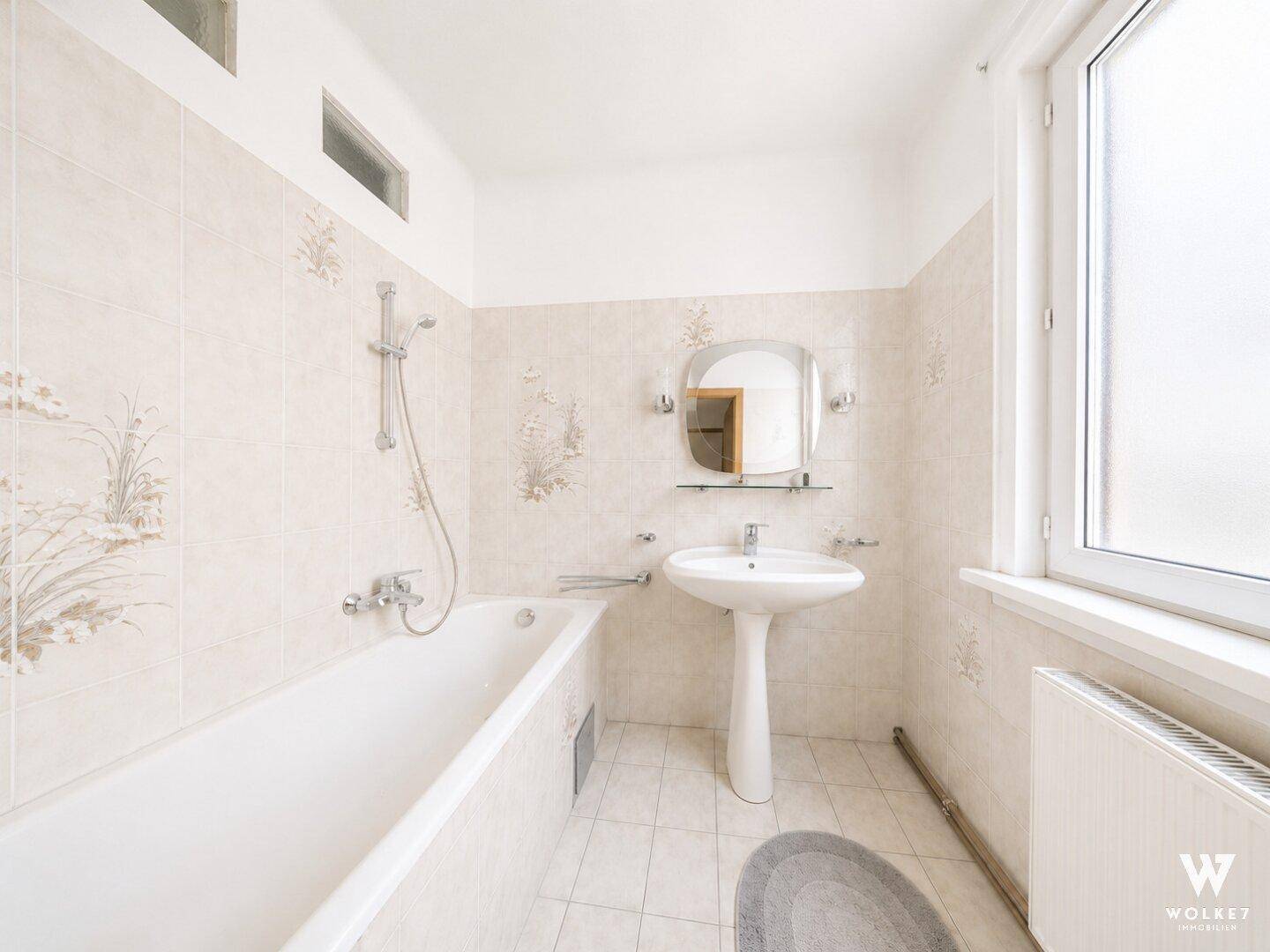Helles Badezimmer mit Badewanne, Standwaschbecken und gemusterten Wandfliesen.