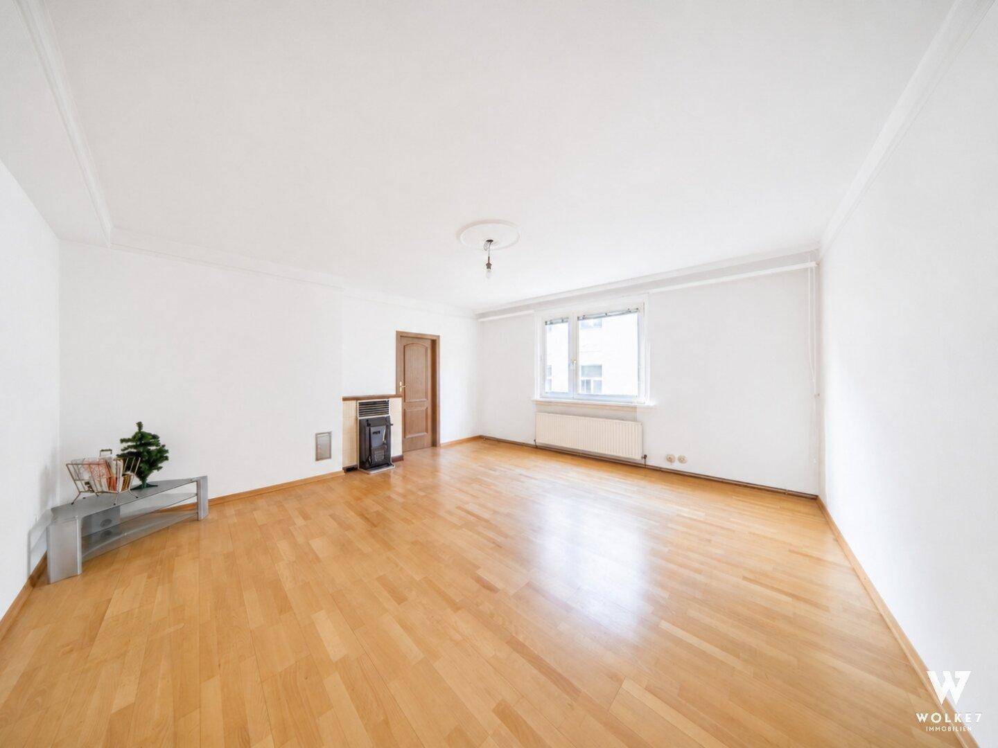 Geräumiges, leeres Wohnzimmer mit hellem Parkettboden und Heizkörper unter dem Fenster.
