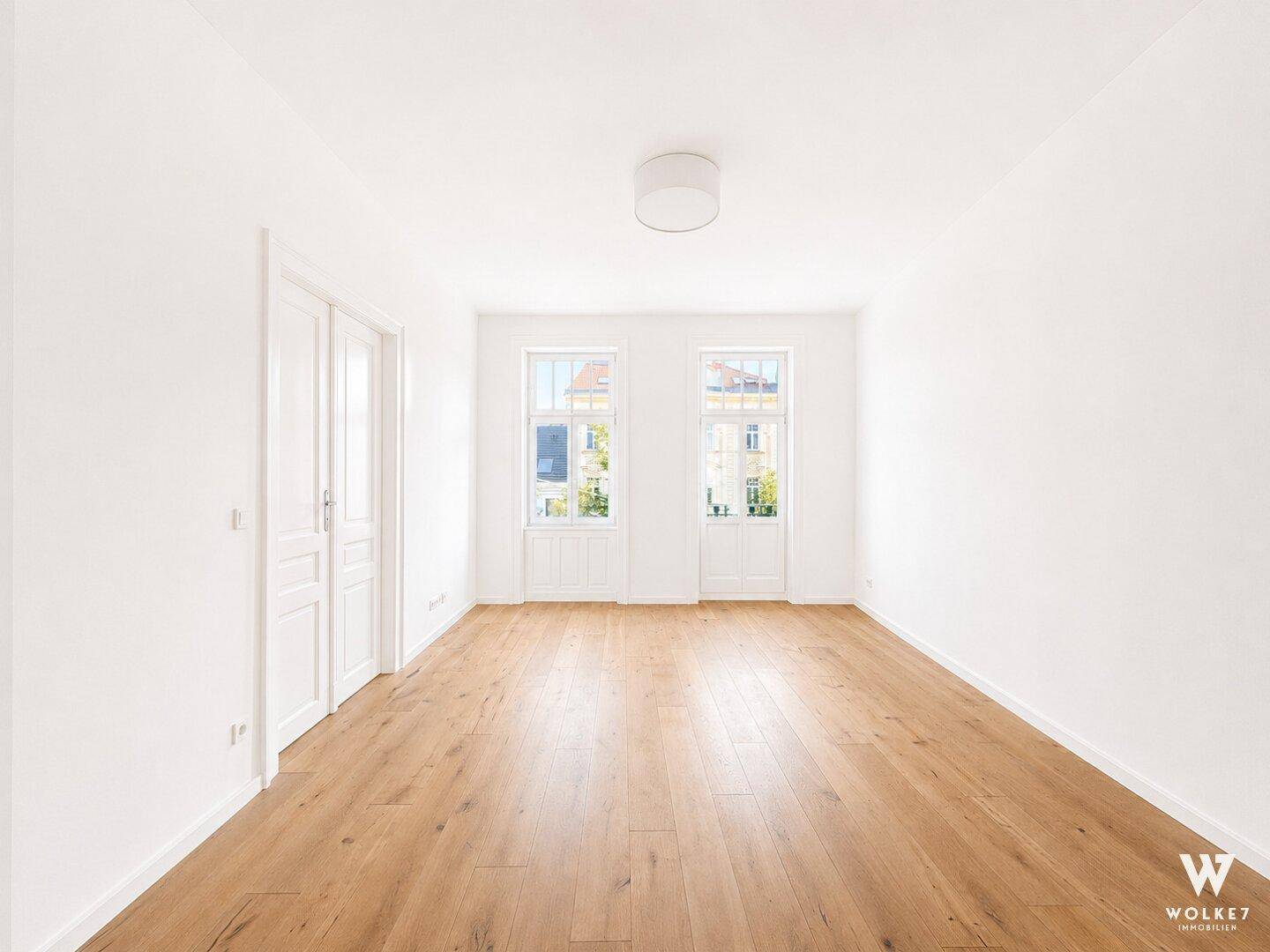 Heller, leerer Raum mit Holzboden, Doppeltüren und zwei hohen Fenstern.