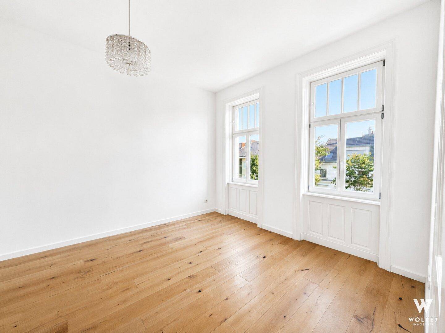 Leerer Raum mit Holzboden, hohen Decken, zwei großen Fenstern und Kronleuchter.