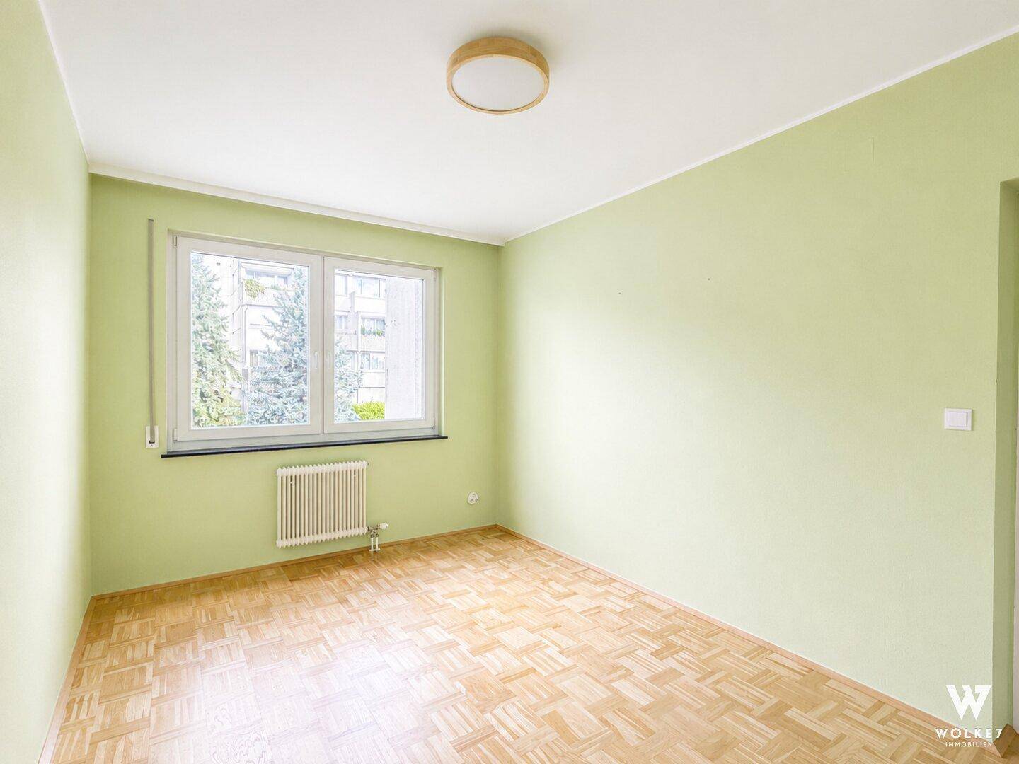Helles, leeres Zimmer mit hellgrünen Wänden, Parkettboden und Blick ins Grüne.