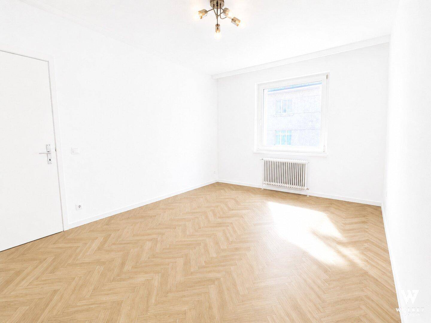 Leeres Schlafzimmer mit hellem Fischgrätparkett, weißen Wänden und einem Fenster für natürliches Licht.