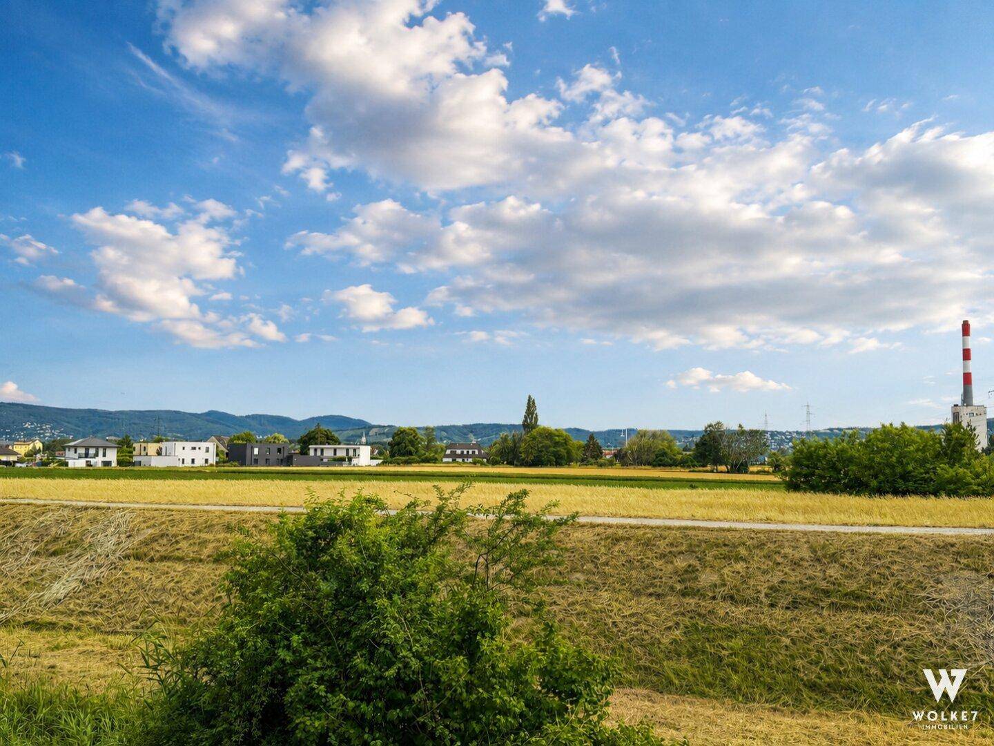 Weitläufige Landschaft mit Feldern, Häusern und einem Industrieschornstein in der Ferne.
