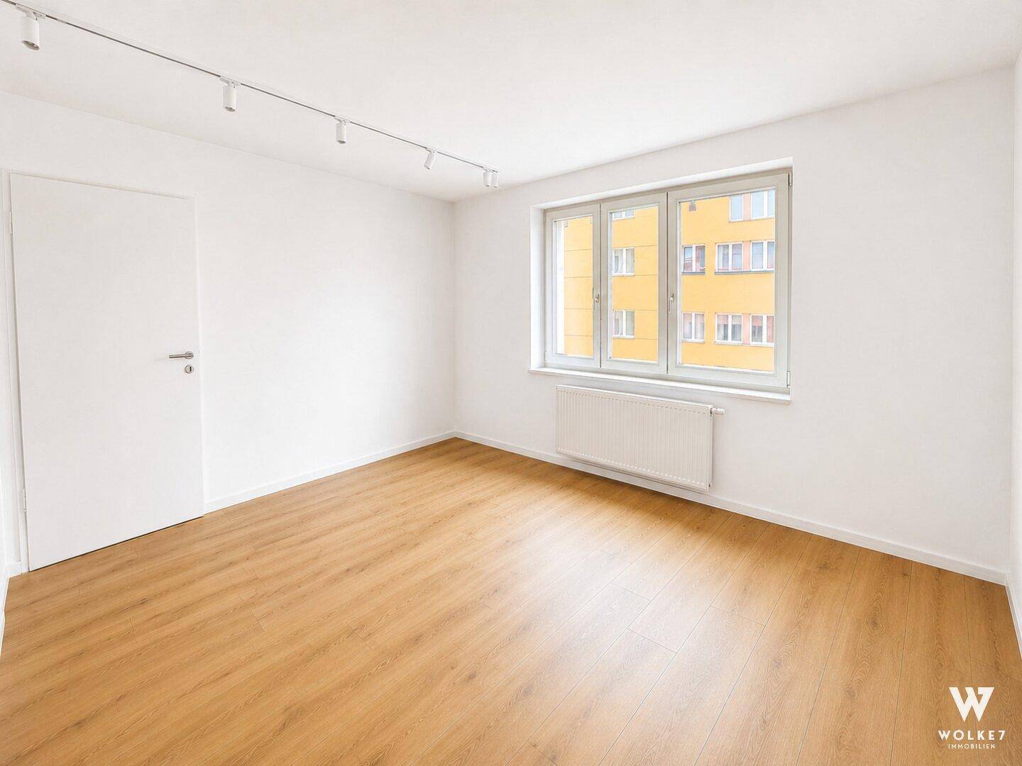 Leeres Zimmer mit Holzfußboden und Fensterblick auf ein gegenüberliegendes Gebäude.