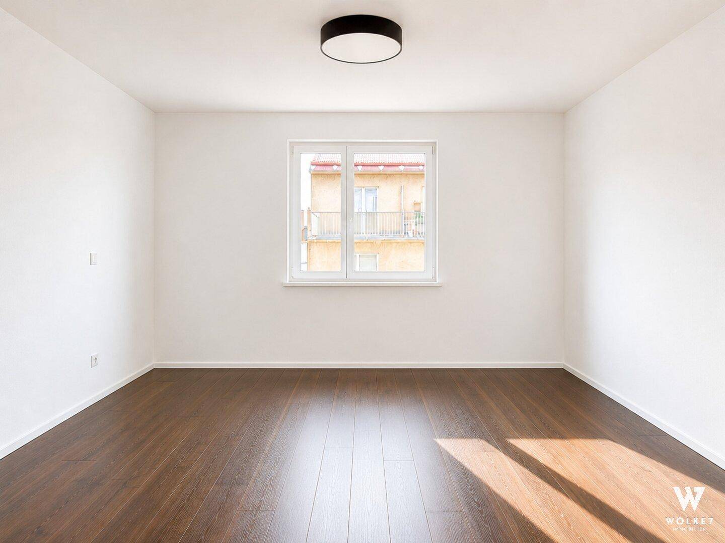 Leerer Raum mit dunklem Holzboden und einem Fenster, das Tageslicht hereinlässt.