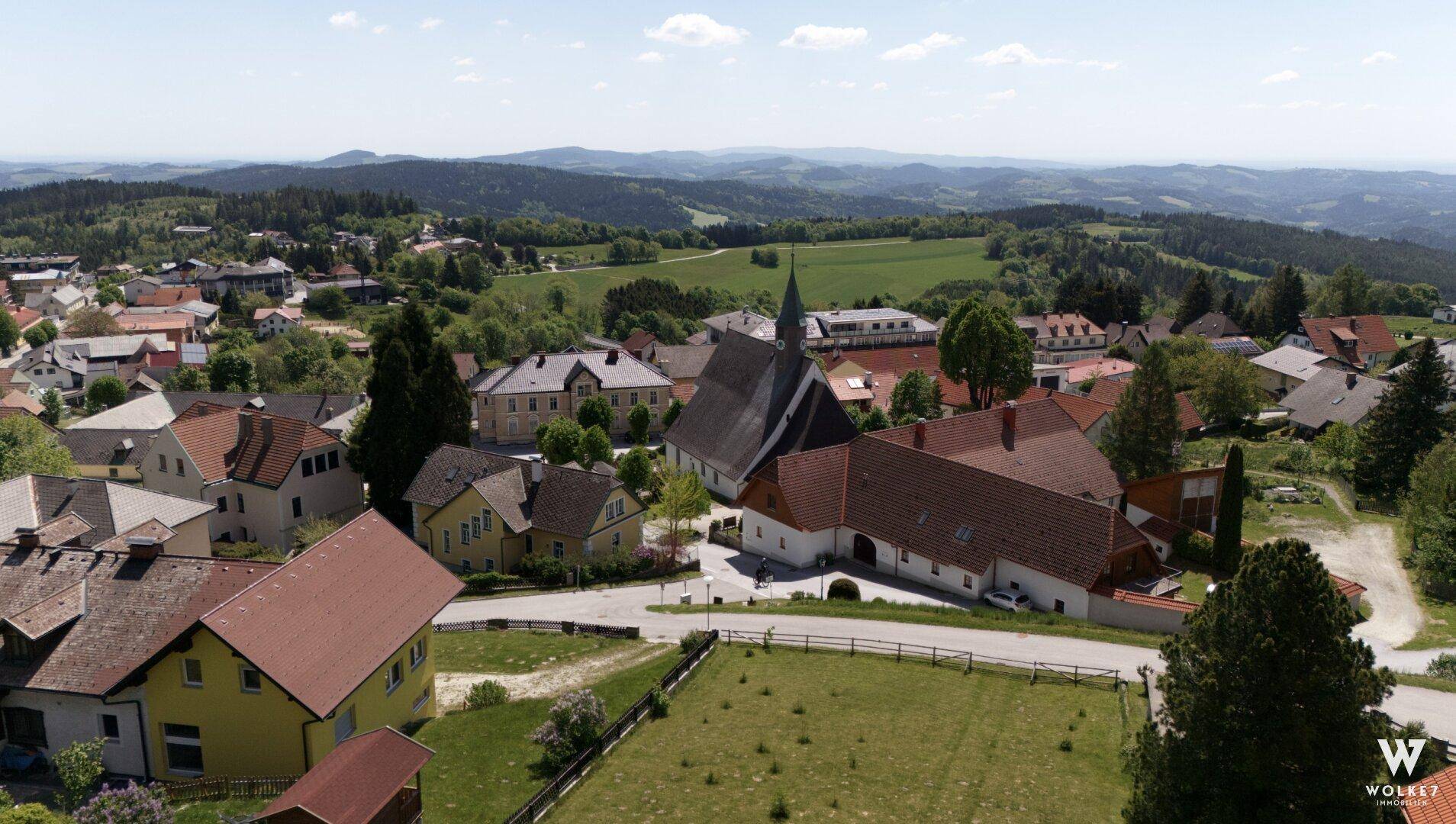 Panoramablick aus der Vogelperspektive auf ein malerisches Dorf, umgeben von sanften Hügeln und Wäldern.