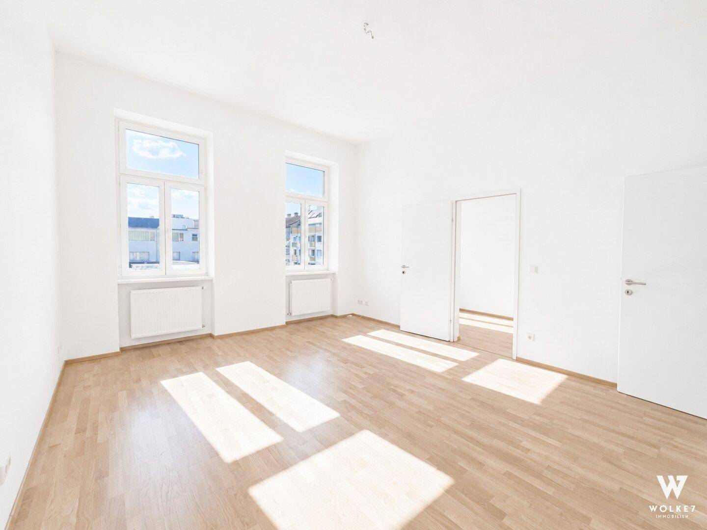 Großes, leeres Zimmer mit hellem Holzboden, weißen Wänden und viel Sonnenlicht durch die Fenster.