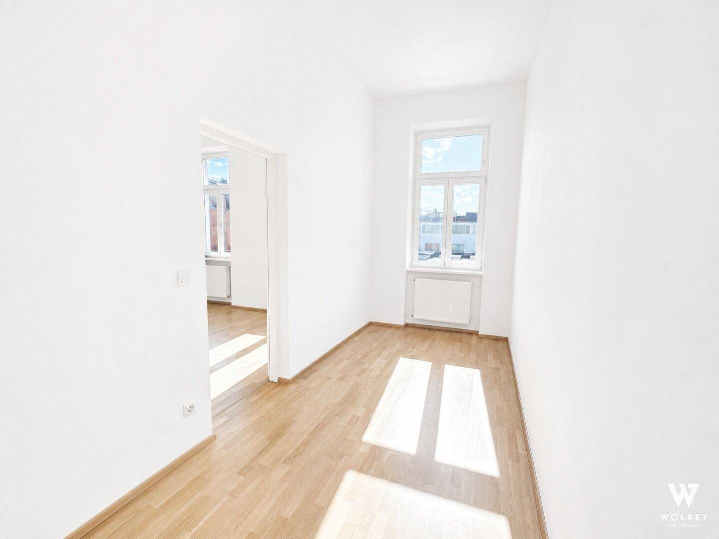 Helles, leeres Zimmer mit Holzboden und Fenster, das zu einem weiteren Raum führt.