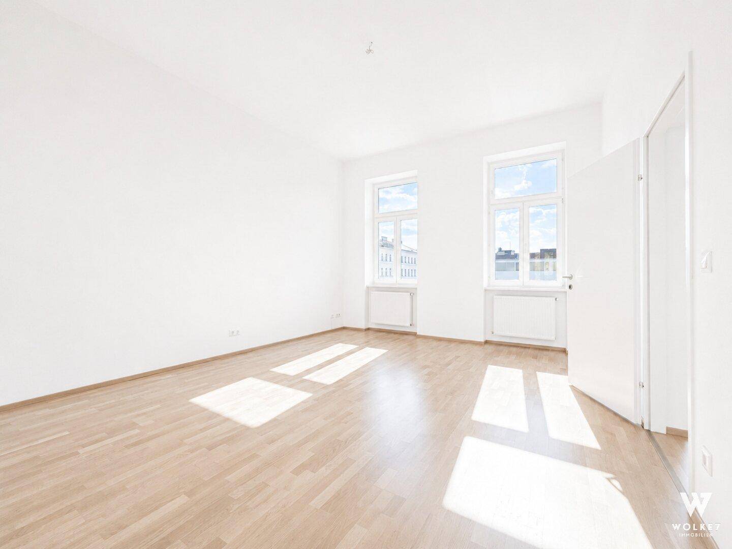 Geräumiges, leeres Zimmer mit hellem Holzboden, weißen Wänden und viel natürlichem Licht durch die Fenster.