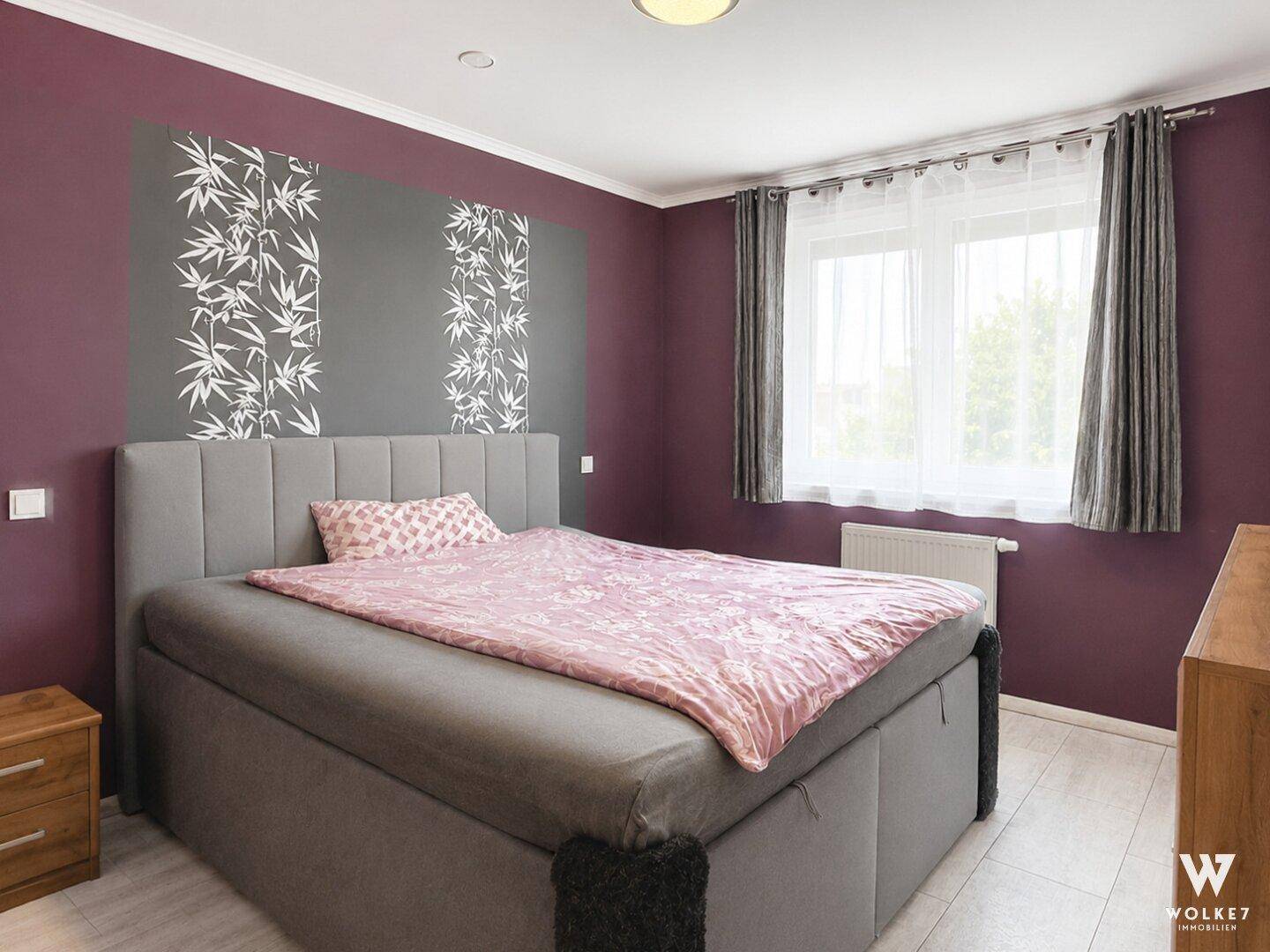 Schlafzimmer mit großem Bett, farbiger Akzentwand und Fenster mit Vorhängen.