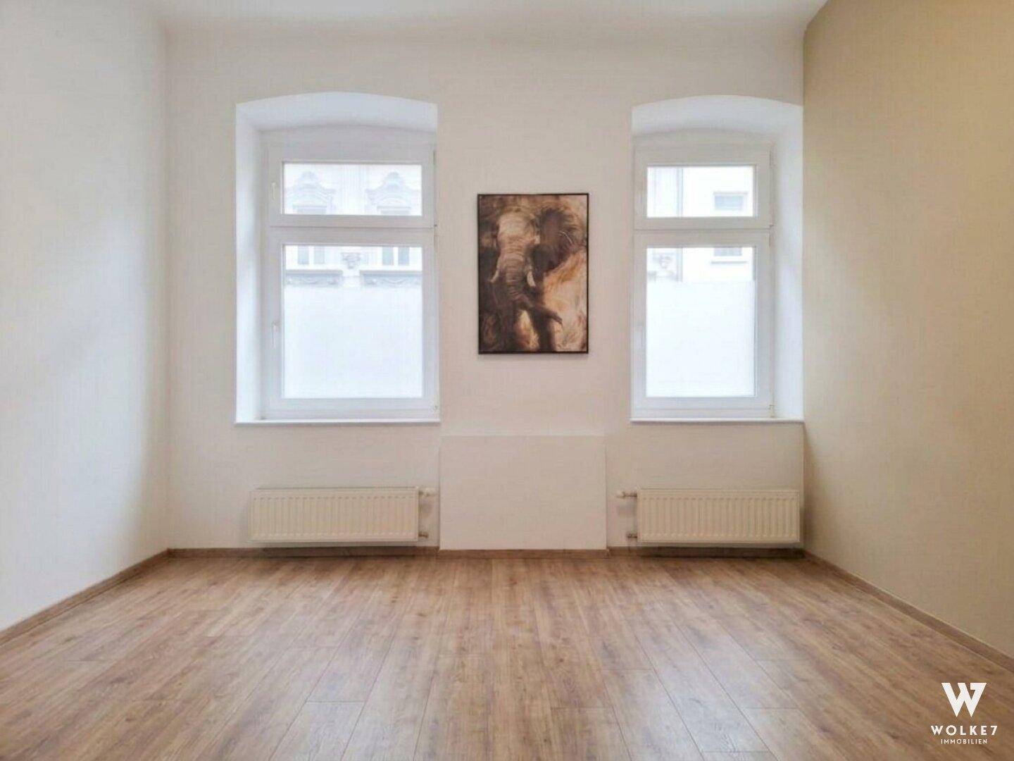 Geräumiges Zimmer mit Holzboden, hellen Wänden und zwei hohen Fenstern.
