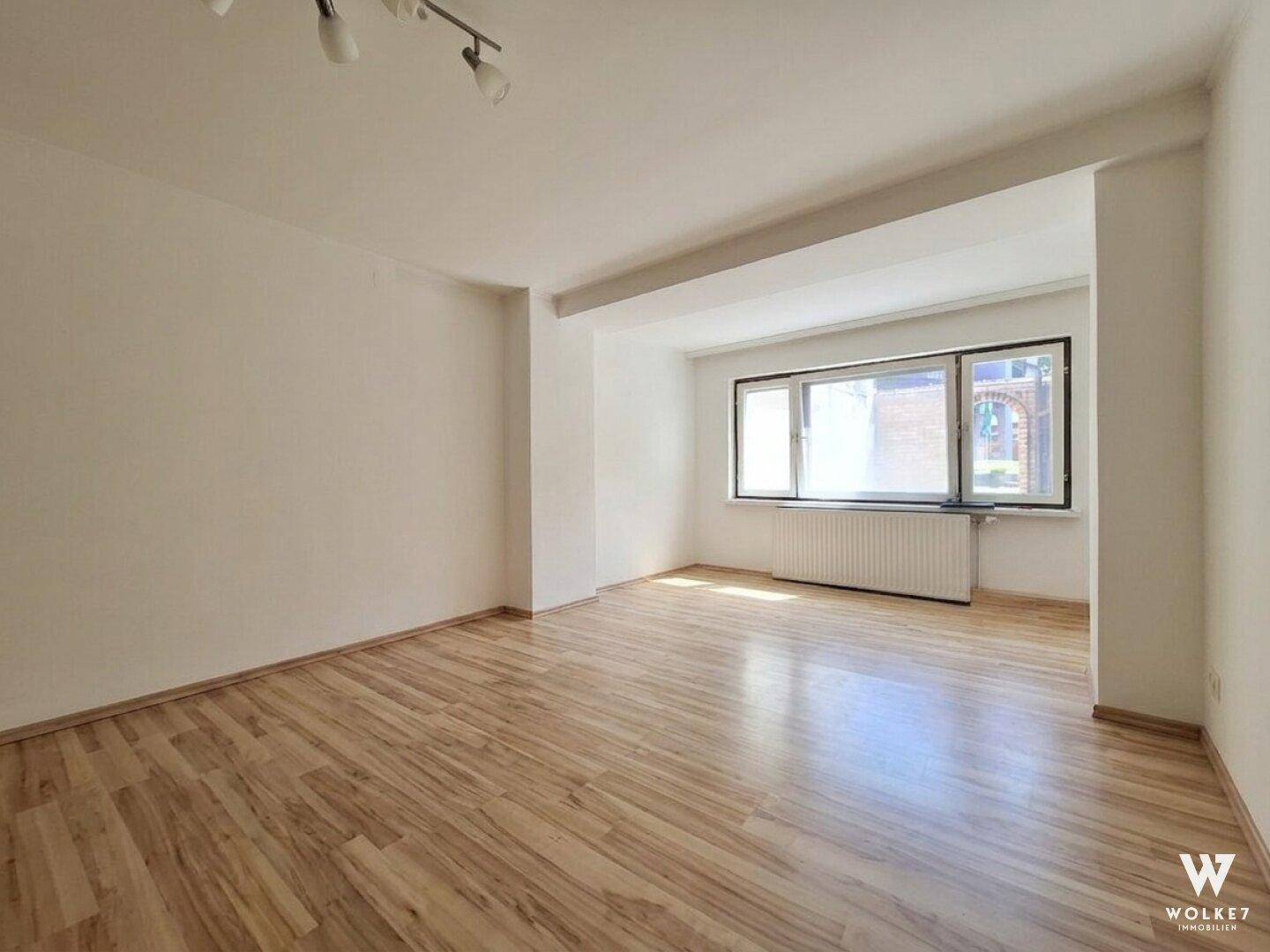 Geräumiger, heller Raum mit Laminatboden und großem Fenster, ideal als Wohnbereich.