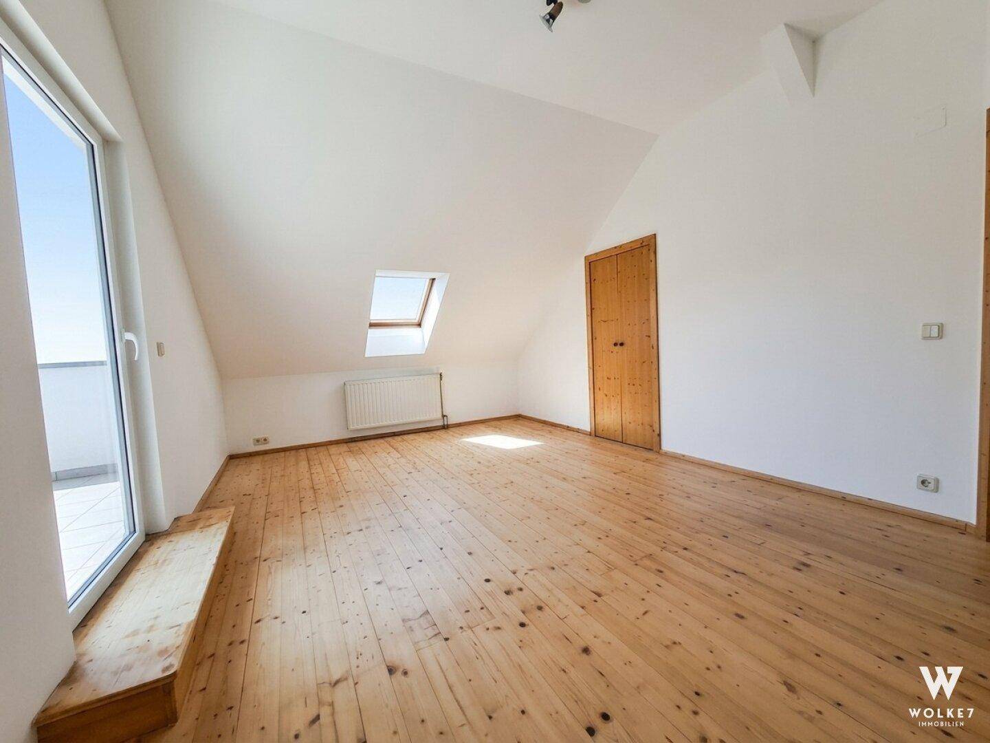 Helles Dachzimmer mit Holzboden, Dachfenster und Zugang zu einem Balkon.