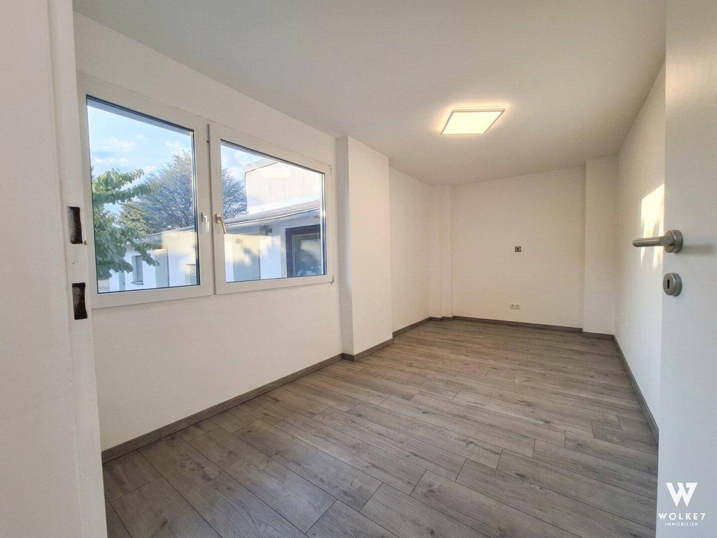 Helles, leeres Zimmer mit großen Fenstern und modernem Laminatboden.