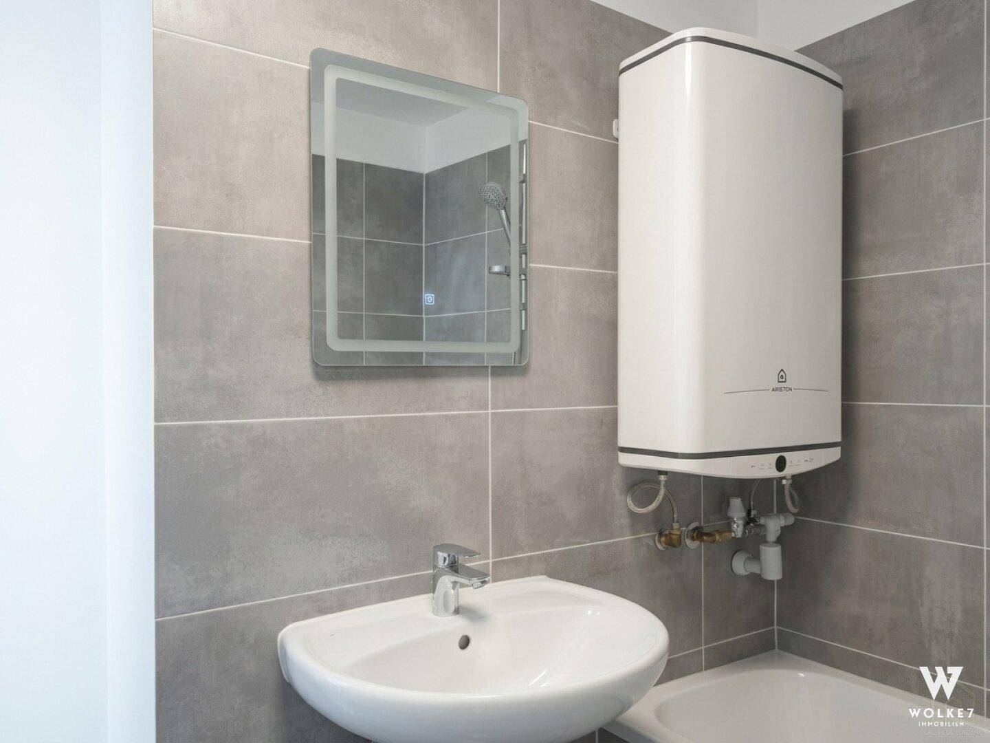 Modernes Badezimmer mit grauen Fliesen, Waschbecken, Spiegel und Warmwasserboiler.