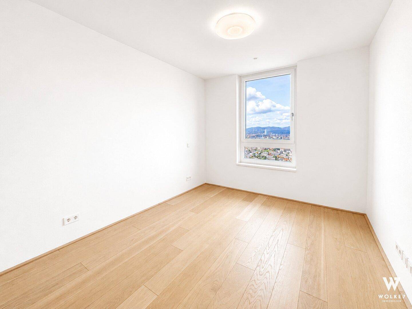 Helles, leeres Schlafzimmer mit Holzboden, großem Fenster und weitem Blick über die Stadt.