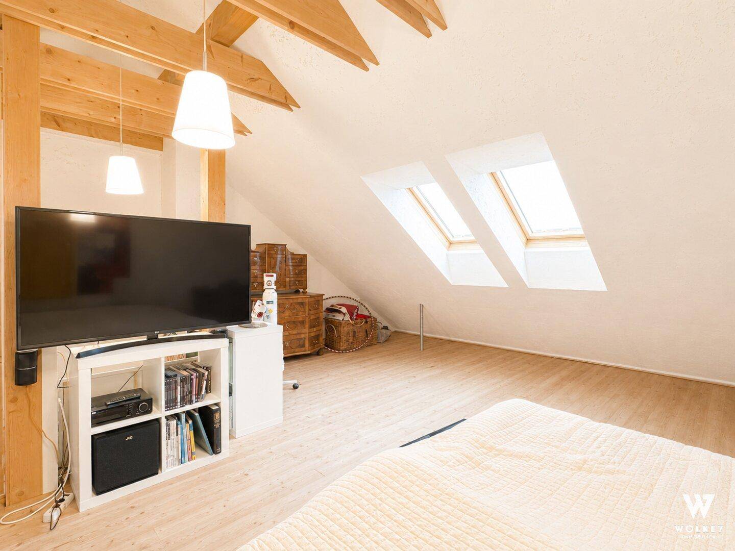 Helles Dachgeschoss-Schlafzimmer mit Holzbalken, Dachfenstern und großem Fernseher.