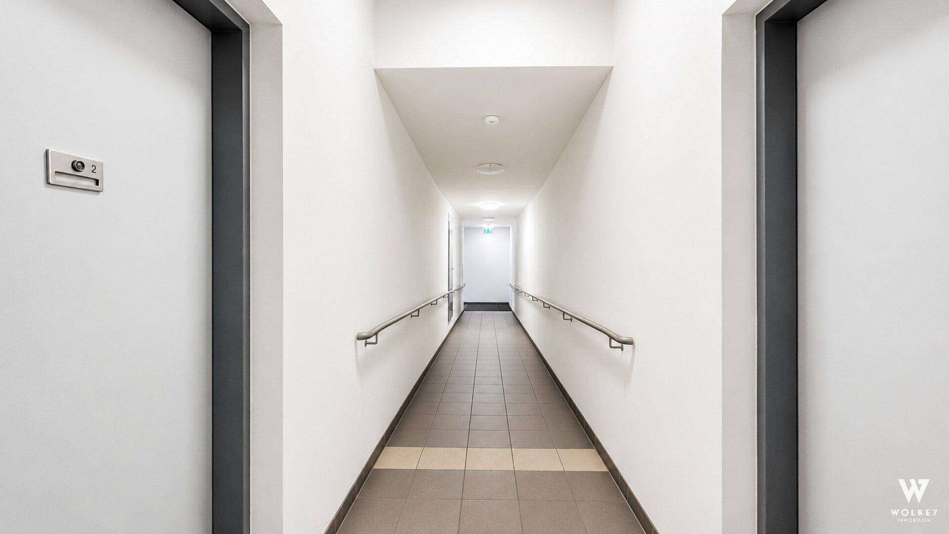 Langer, heller Flur mit Handläufen und Fliesenboden in einem Mehrfamilienhaus.