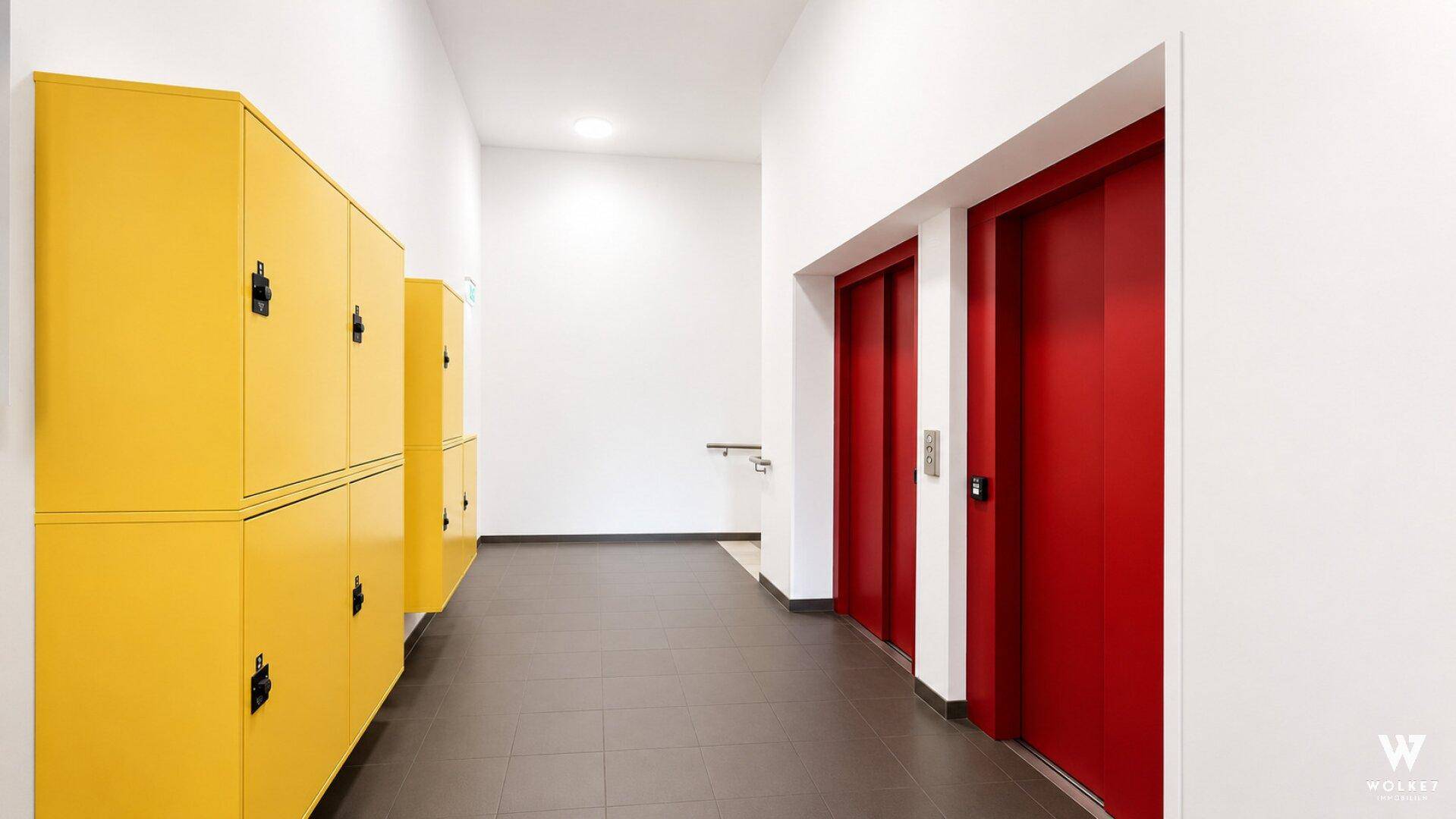 Heller Flur mit roten Aufzugtüren und gelben Schließfächern in einem modernen Gebäude.