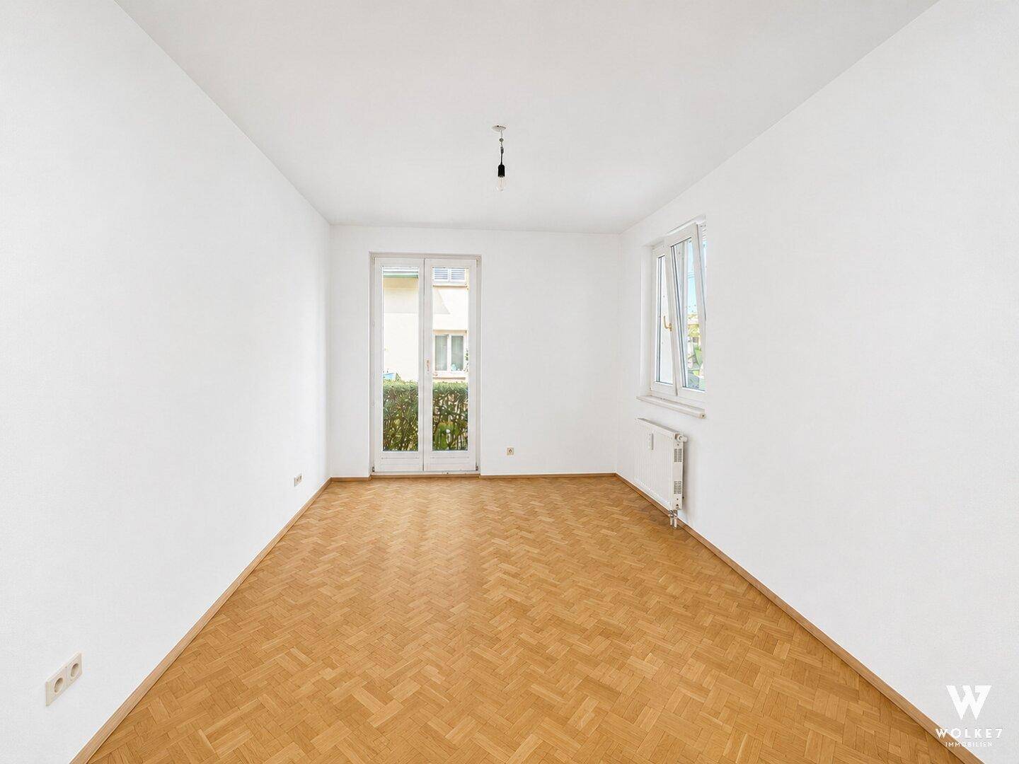 Leeres Zimmer mit Parkettboden und Fenstertür, die viel Tageslicht hereinlässt.
