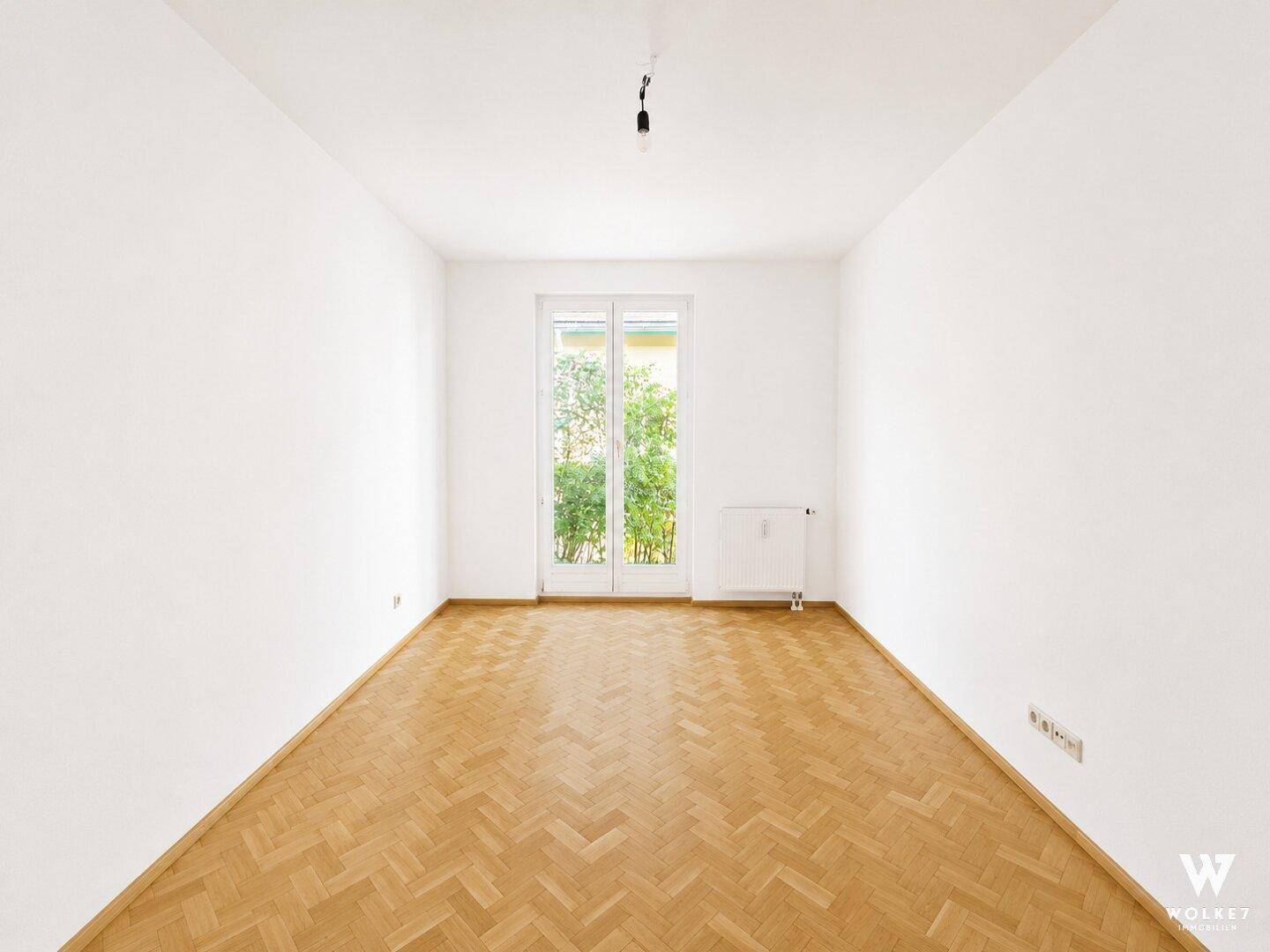 Helles, leeres Zimmer mit Fischgrätparkett und Zugang zu einem Außenbereich.