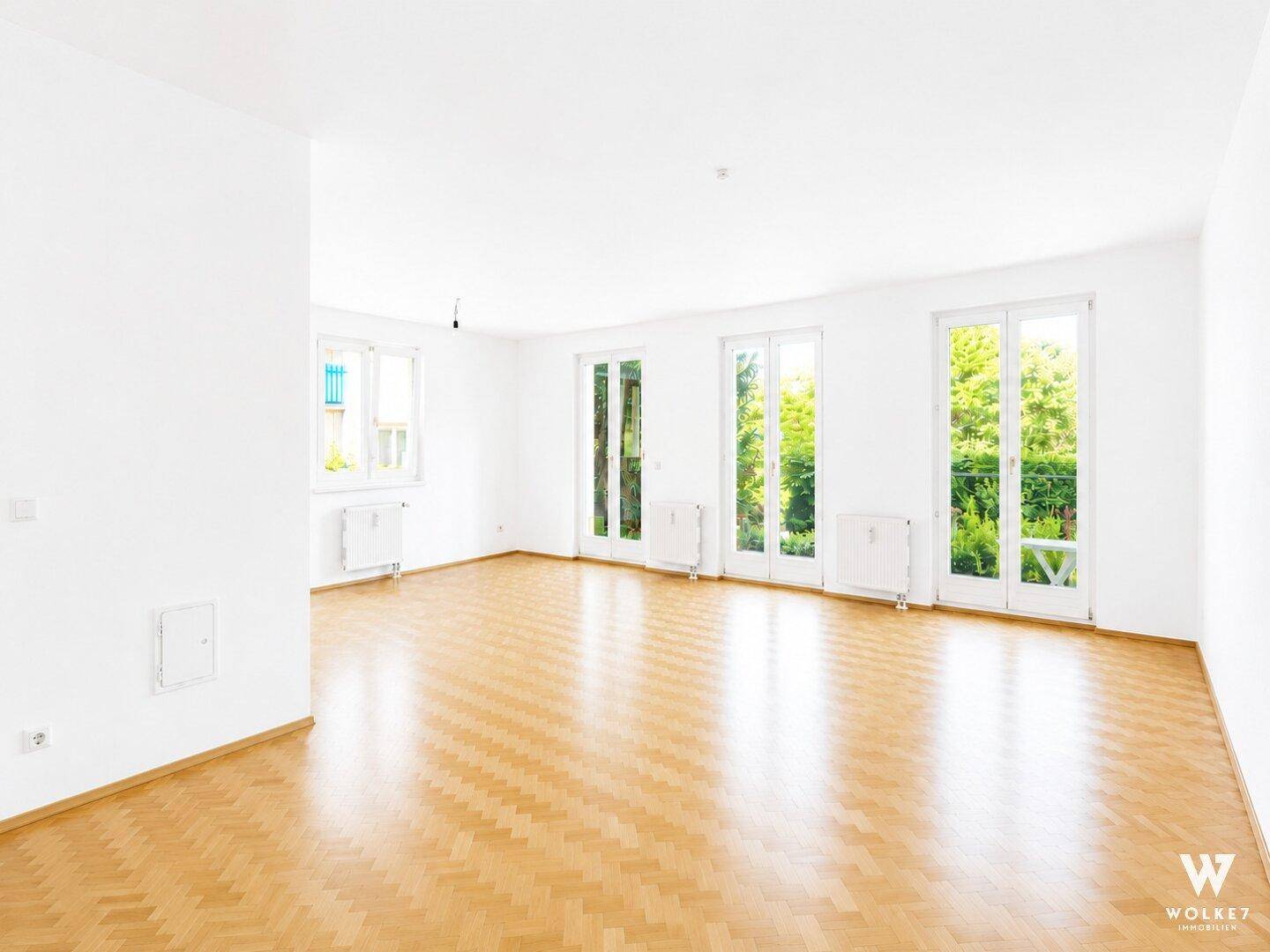 Geräumiges, leeres Wohnzimmer mit Fischgrätparkett und großen Fenstertüren zum Garten.