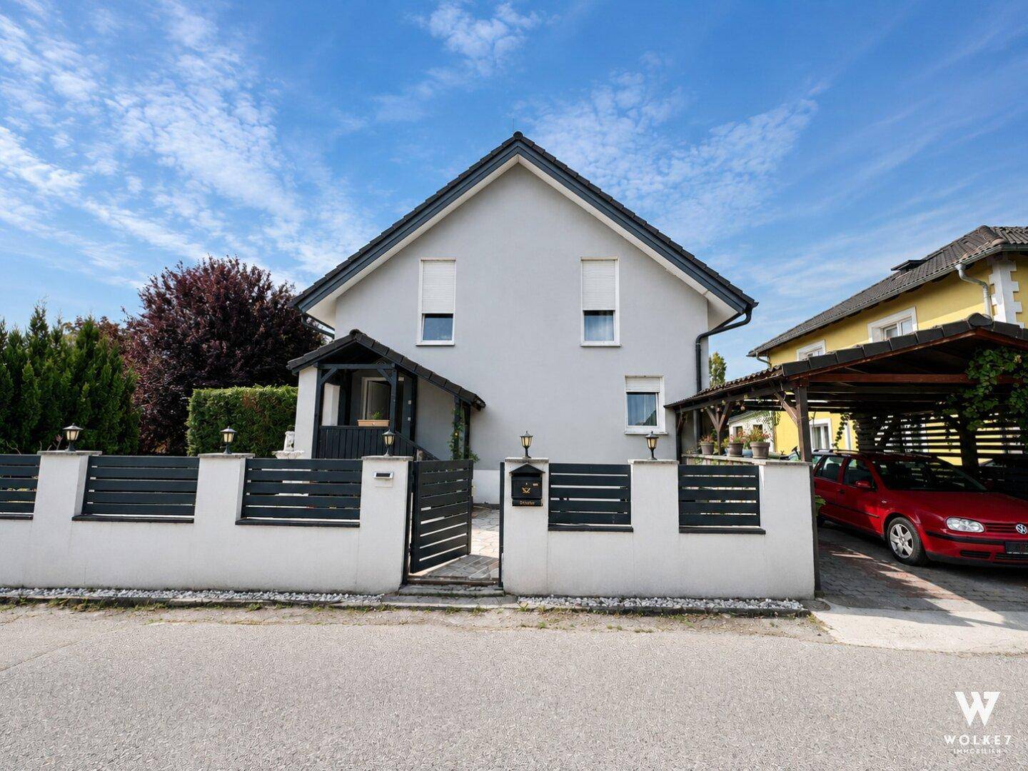 Moderne Hausfassade mit Zaun, Eingangsbereich und einem Carport für Fahrzeuge.