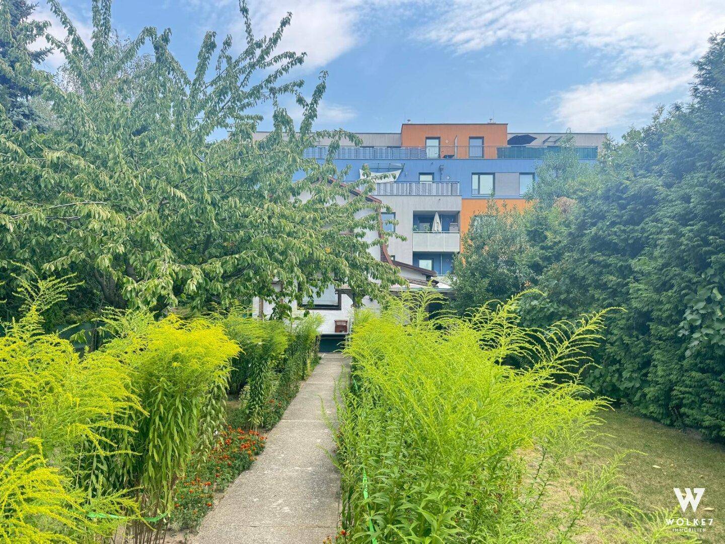 Gartenweg umgeben von üppigem Grün führt zu einem modernen Mehrfamilienhaus mit farbiger Fassade.