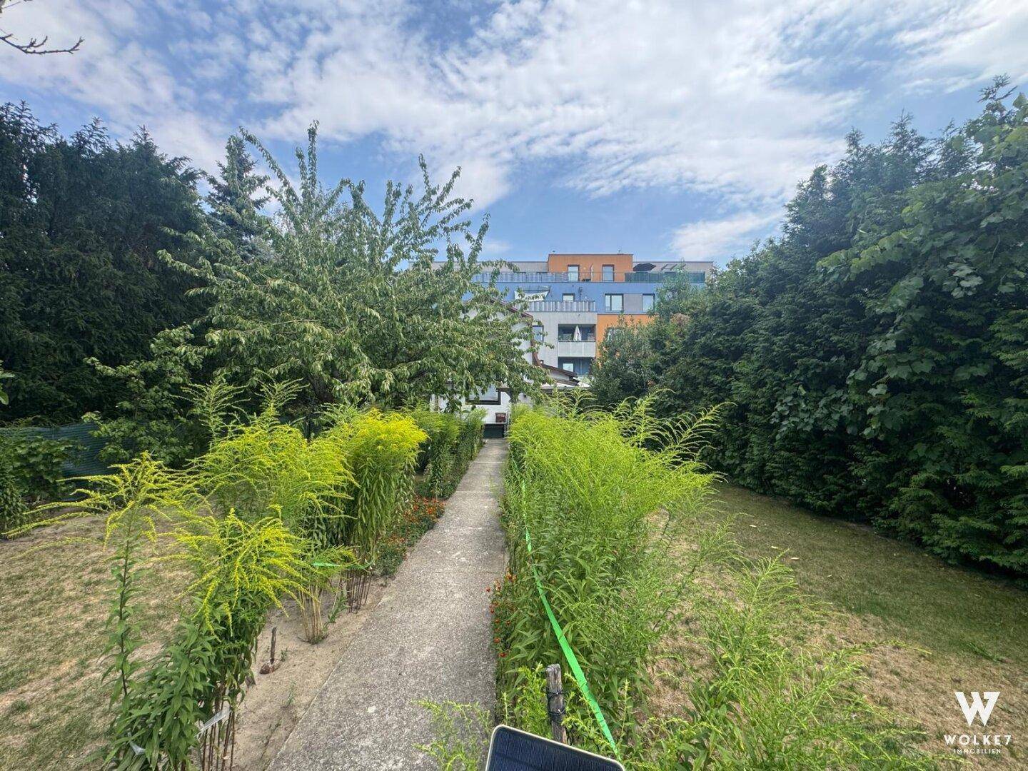 Langer Gartenweg durch grüne Bepflanzung mit Blick auf ein modernes Gebäude unter blauem Himmel.