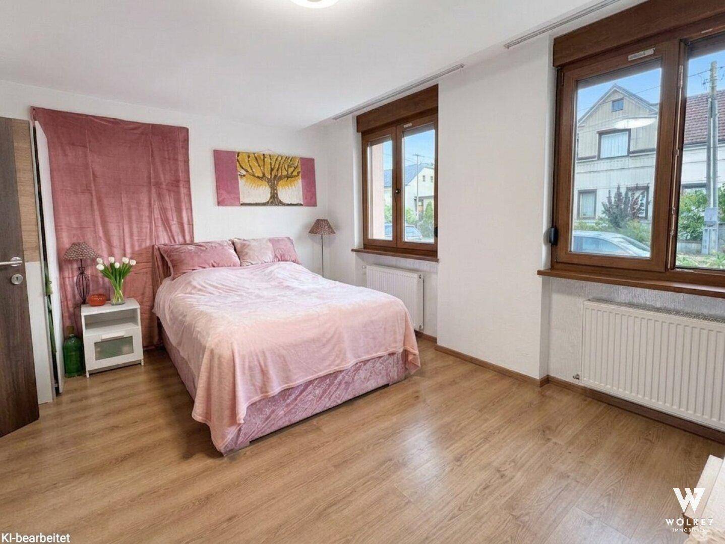 Helles Schlafzimmer mit rosa Bettwäsche, Laminatboden und zwei Fenstern mit Blick nach draußen.