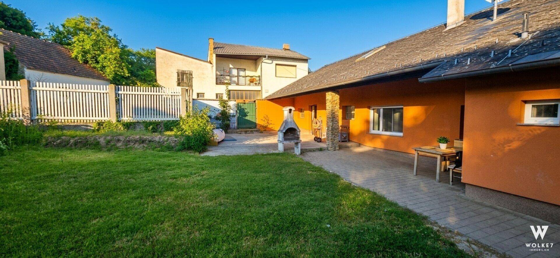 Sonniger Gartenbereich mit Rasen, gepflasterter Terrasse und festem Grill hinter dem Haus.