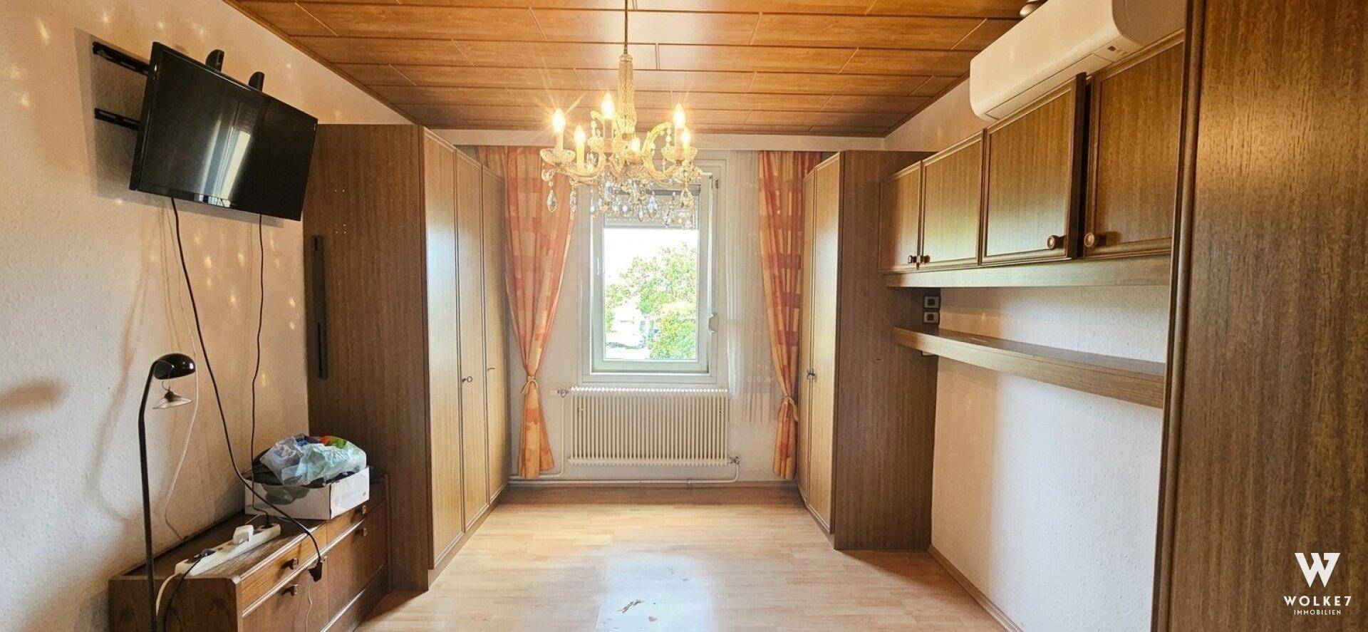 Schlafzimmer mit Holzdecke, Einbauschränken und Kronleuchter, hell durch das Fenster.
