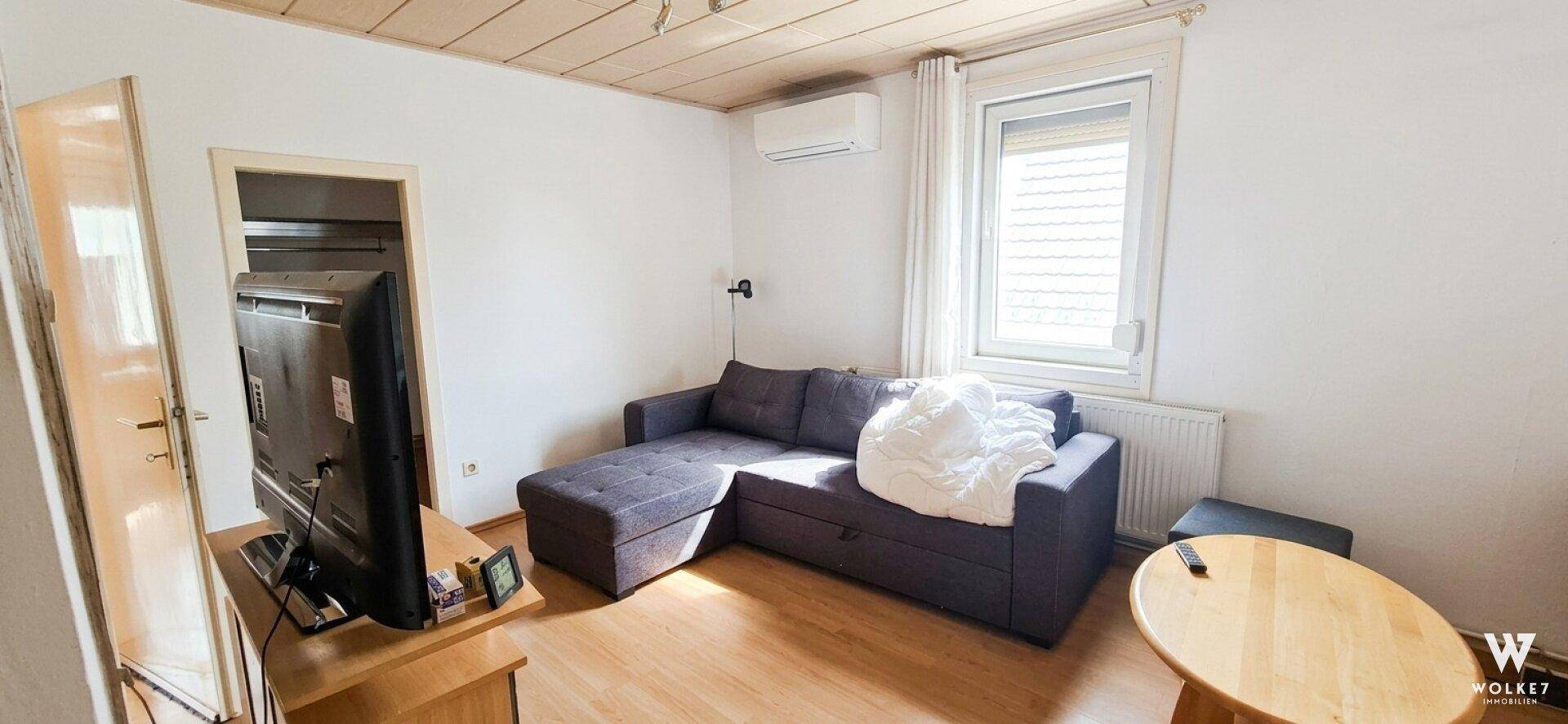 Kleines Wohnzimmer mit Sofa, Fernseher und Klimaanlage, hell durch das Fenster.