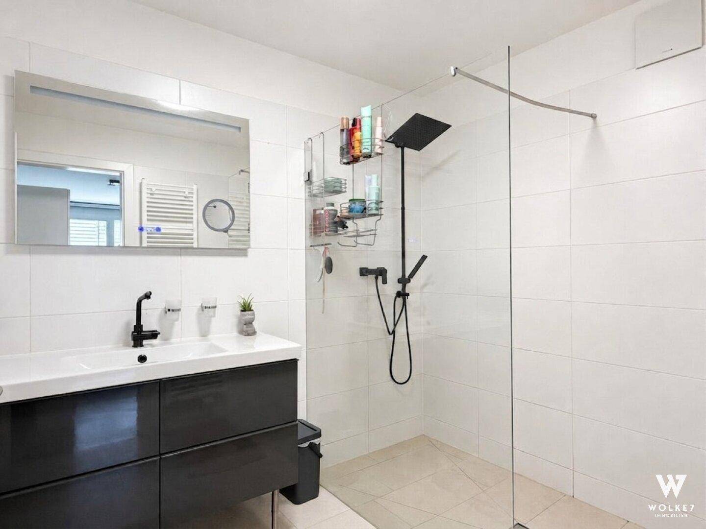 Modernes Badezimmer mit großer begehbarer Dusche, weißen Fliesen und dunklem Waschtischunterschrank.