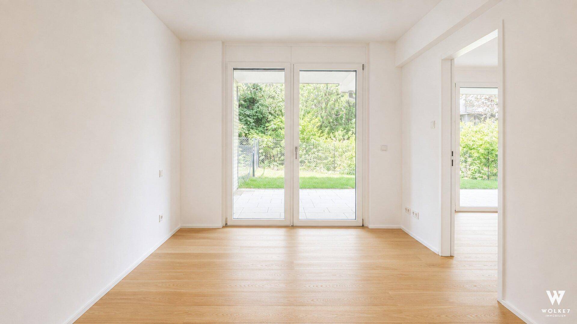 Geräumiger, heller Raum mit Holzboden und großen Fenstertüren, die den Blick ins Grüne freigeben.