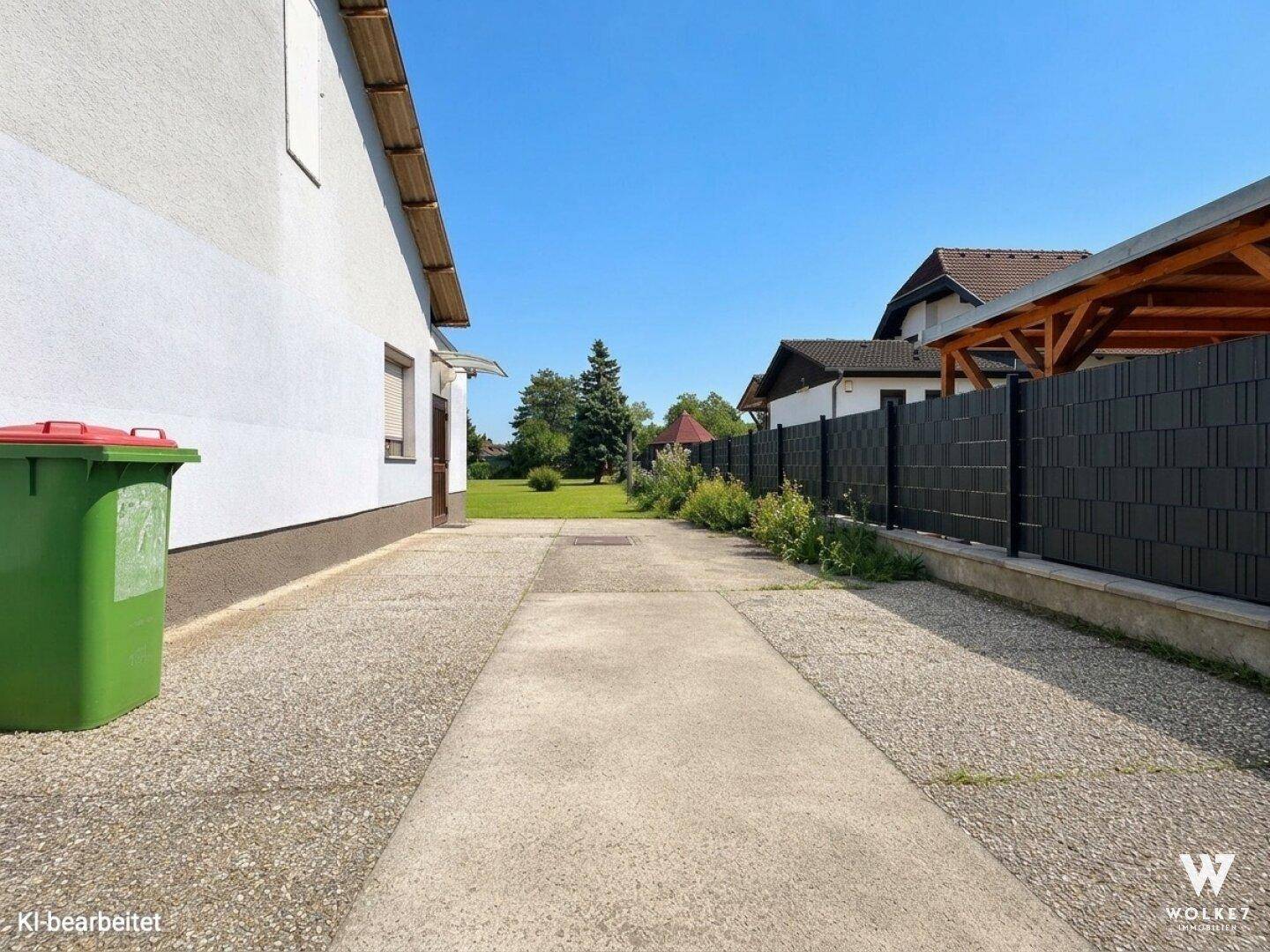 Zufahrtsweg zwischen Gebäuden mit einem grünen Mülleimer und einem modernen Zaun.