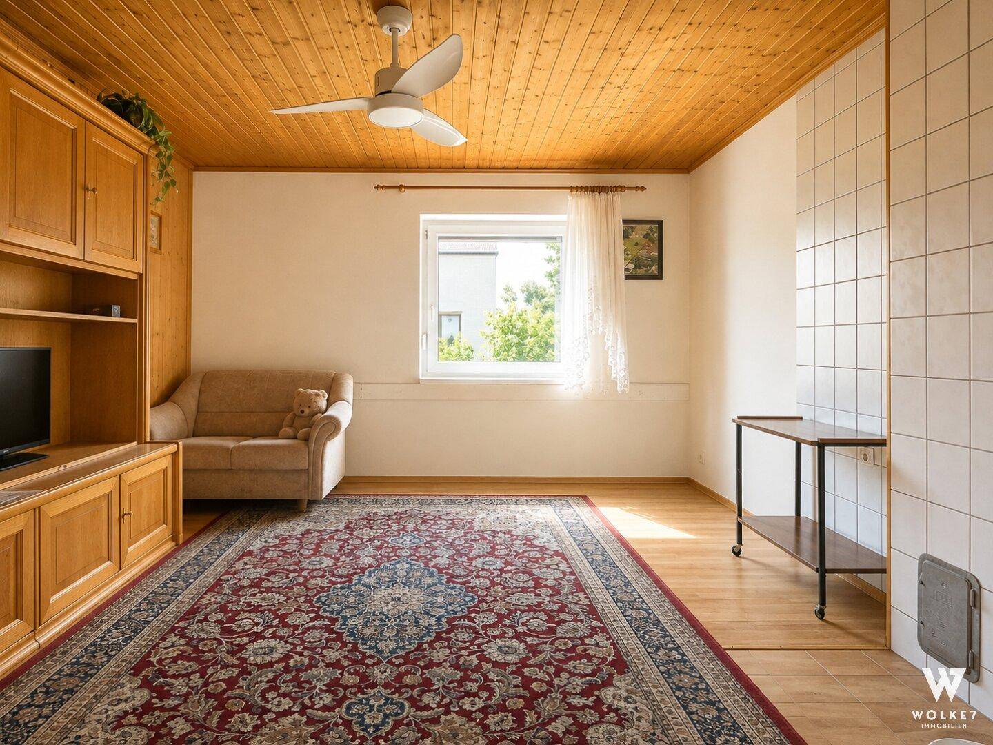 Helles Wohnzimmer mit Holzbalkendecke, großem Teppich und Kachelofen.