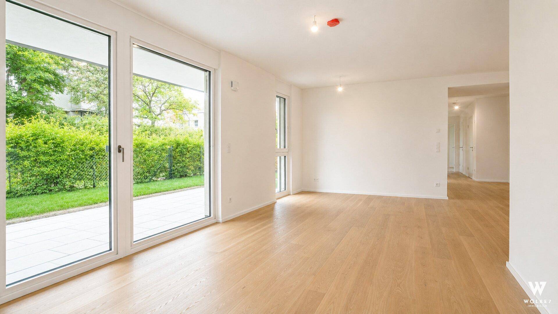 Heller, leerer Wohnraum mit großen Fenstern und Zugang zum Garten, ausgestattet mit Holzparkettboden.