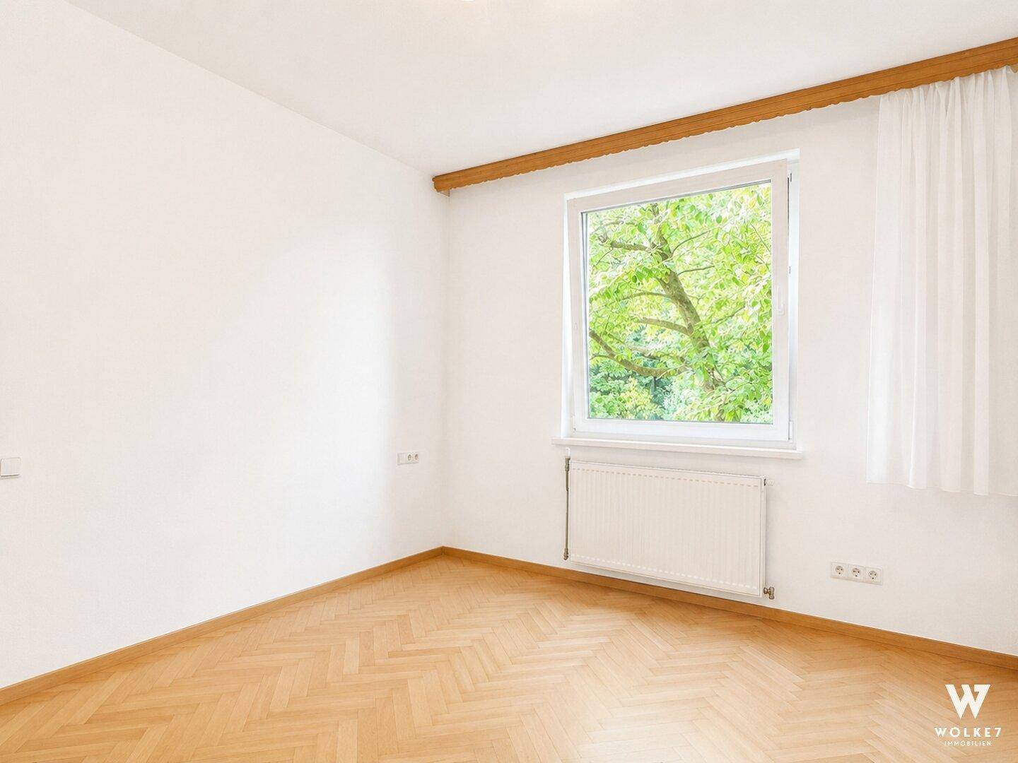 Heller, leerer Raum mit Fischgrätparkett und Fenster mit Ausblick auf grüne Bäume.