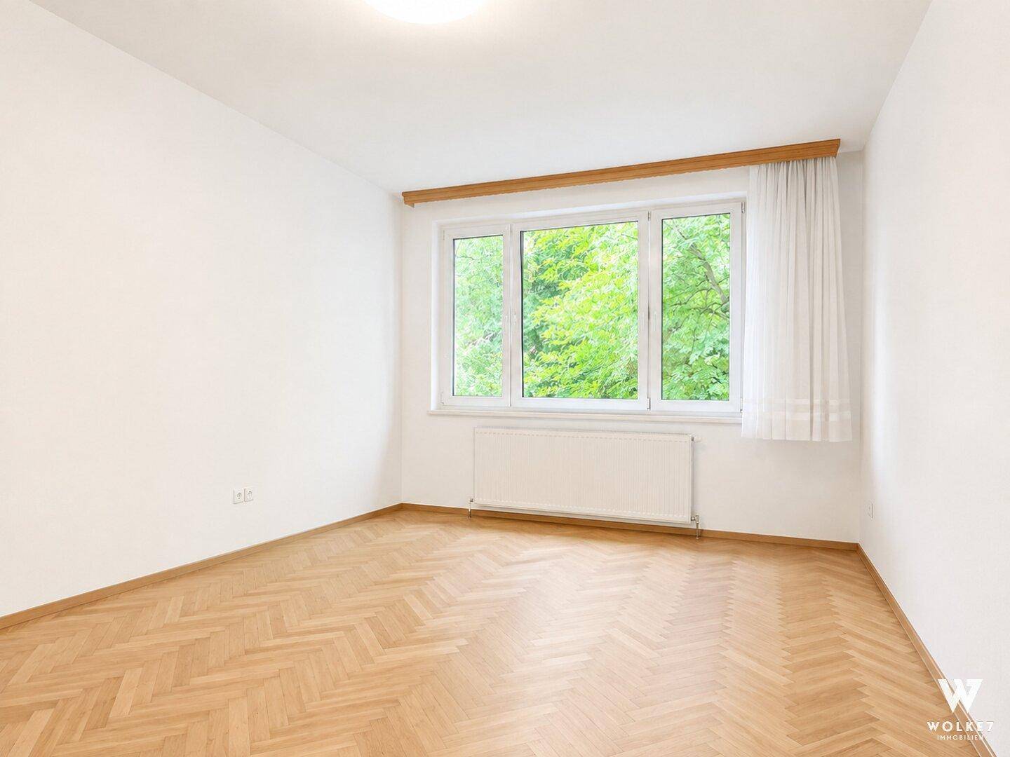 Leerer Raum mit hellem Fischgrätparkett und großem Fenster mit Blick ins Grüne.